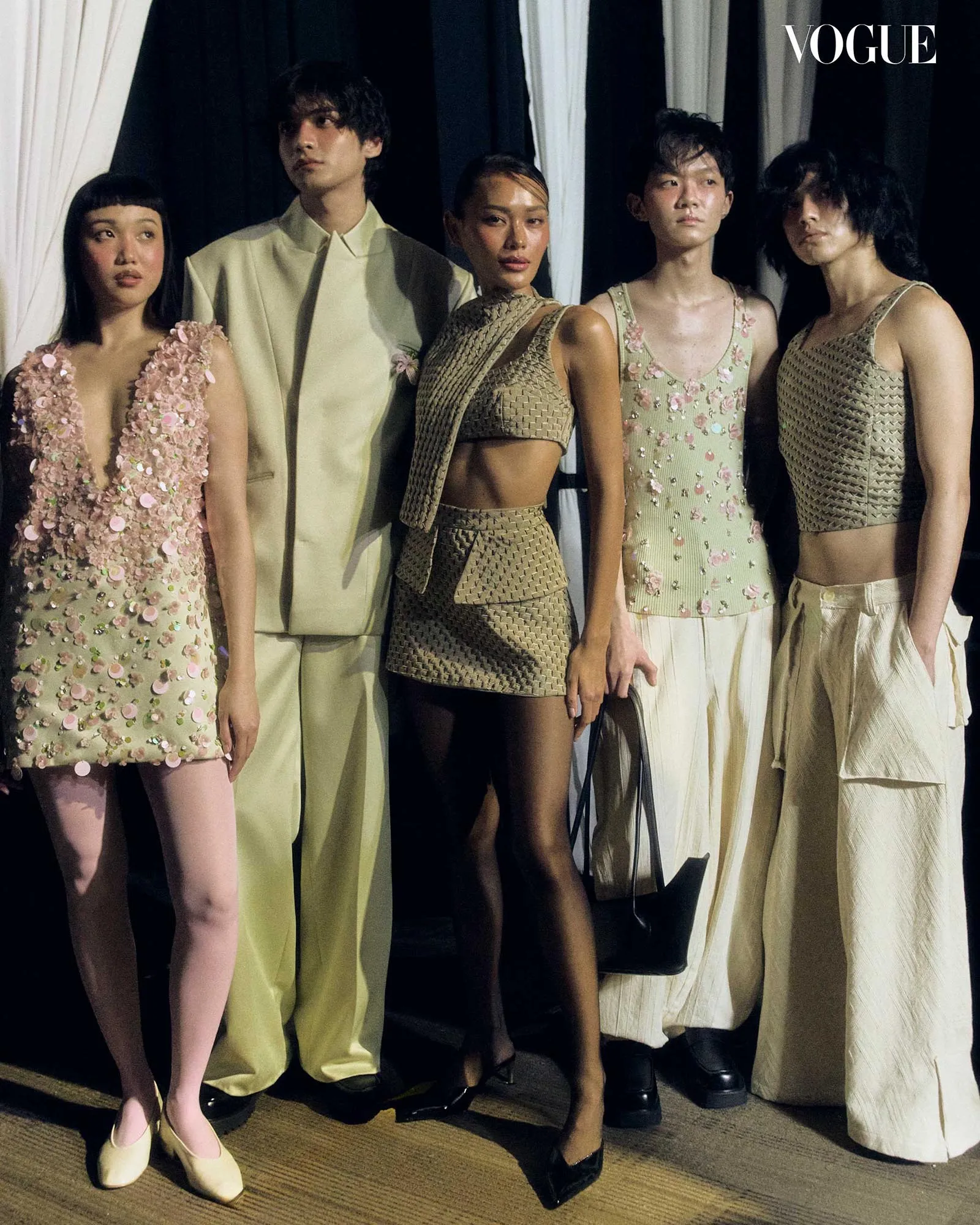 Group of five fashion models in coordinated beige and pastel outfits posing backstage; Vogue logo visible in the corner.