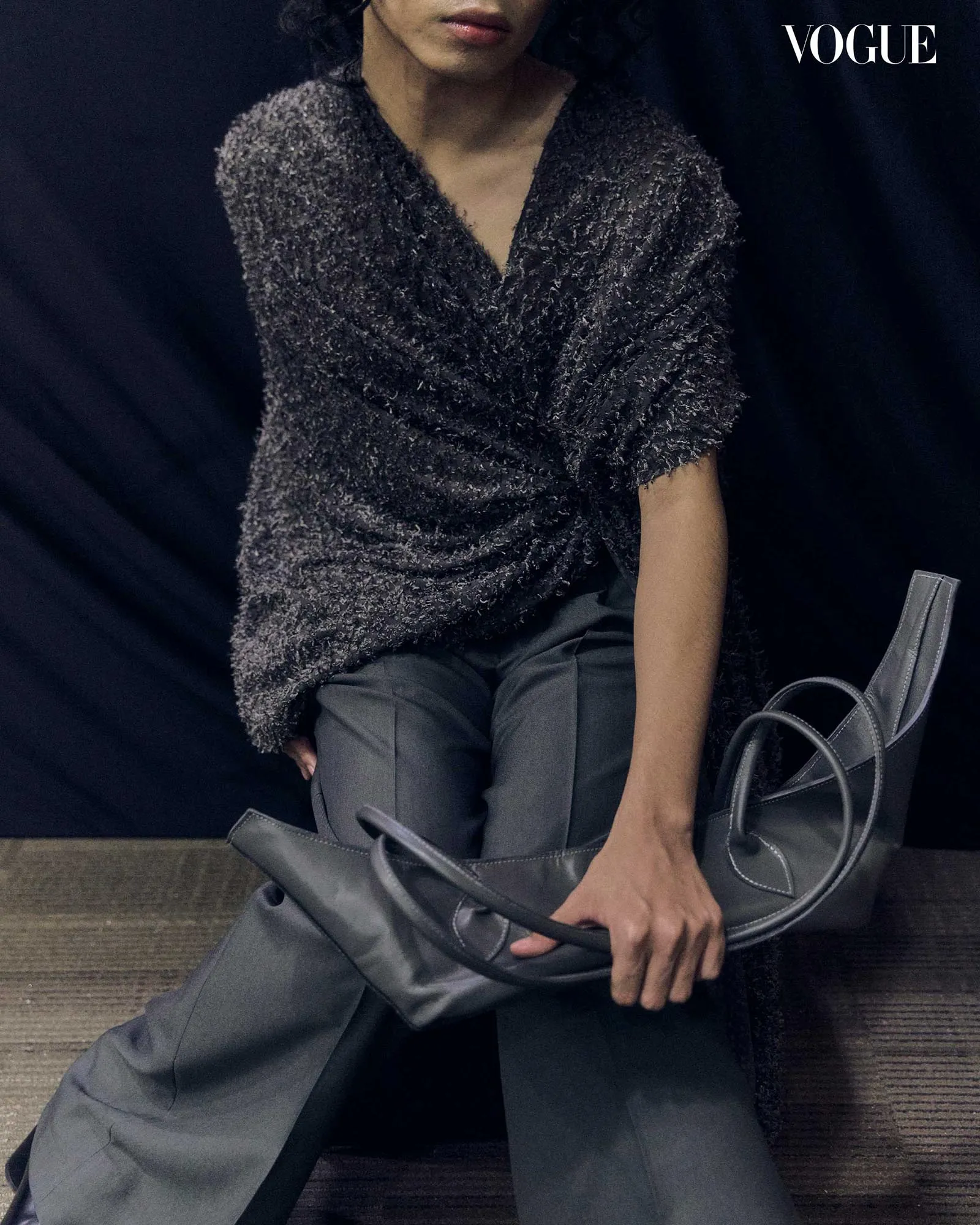 Model seated in a dark, textured wrap top and gray pants, holding a large gray handbag; Vogue logo in the top-right corner.