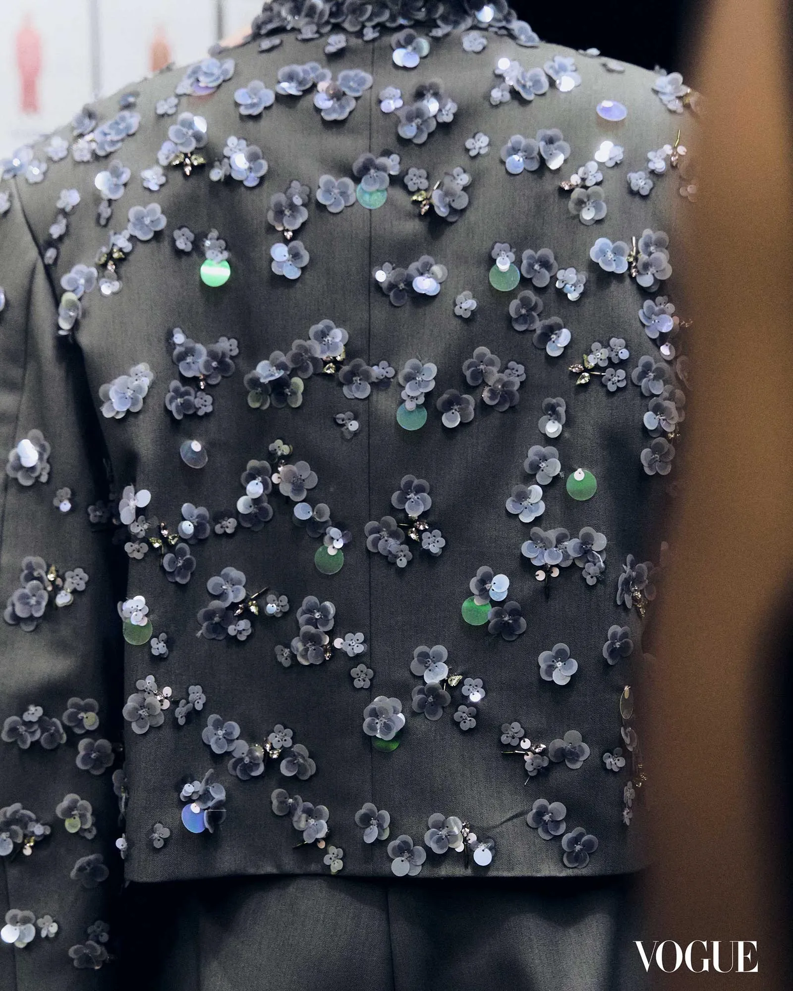 Close-up of a dark fabric dress covered with small translucent gray flower appliqués; Vogue watermark in the bottom-right corner.