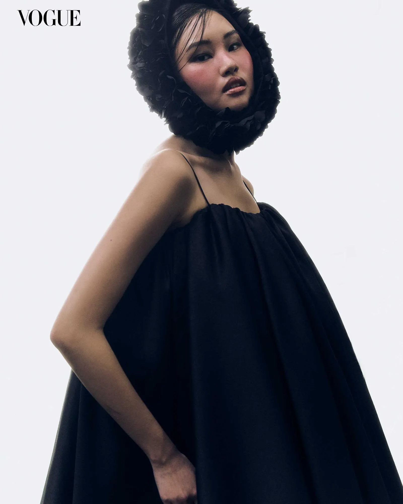 Vogue logo in the top-left corner; model in a black strapless dress with a voluminous black feathered hood against a pale background.