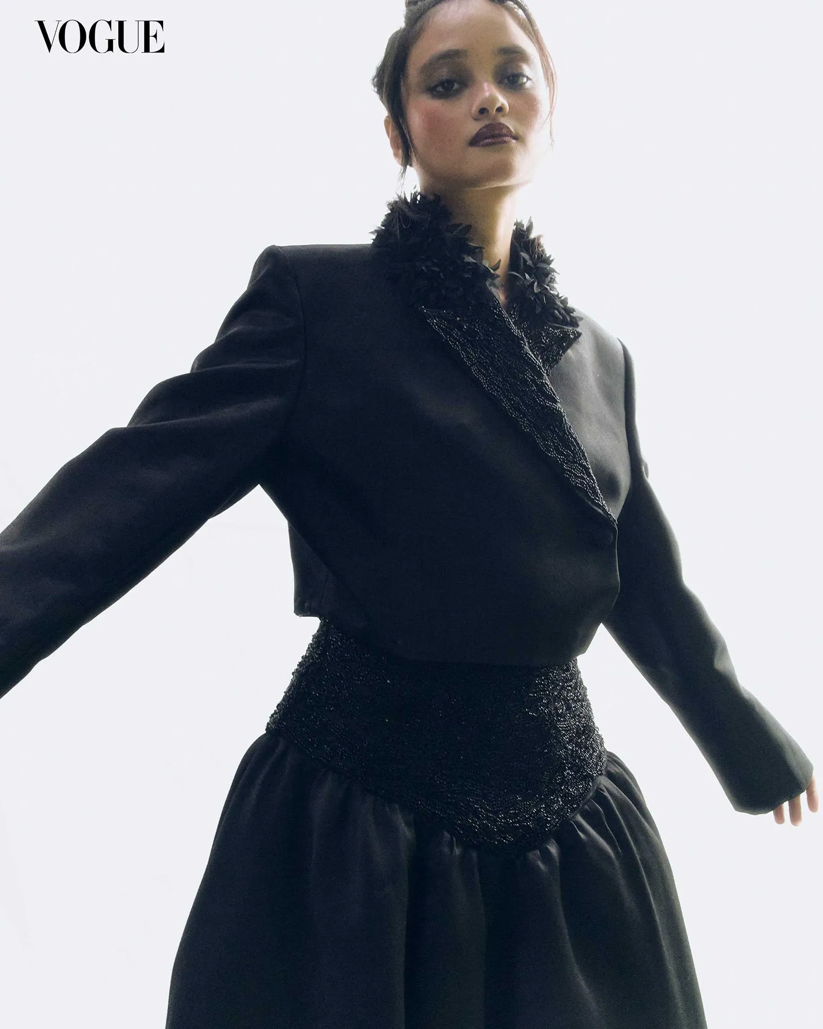Female model wearing a black tailored blazer with feathered lapels and a glittery belt, posed against a pale background with Vogue logo in the corner.