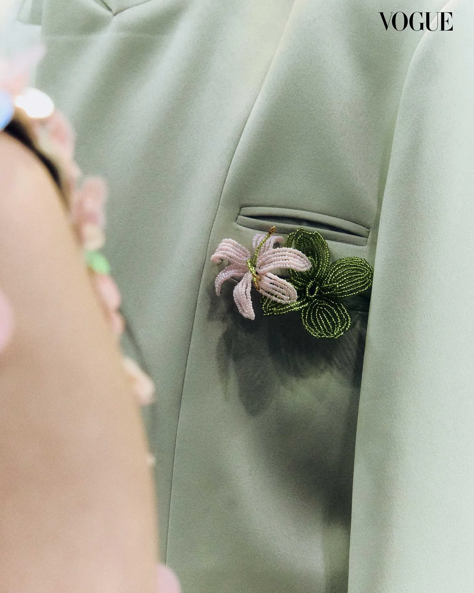 Close-up of a pale mint blazer with a beaded pink and green floral corsage in the chest pocket; partial view of a person in the foreground and the Vogue logo in the top-right corner.