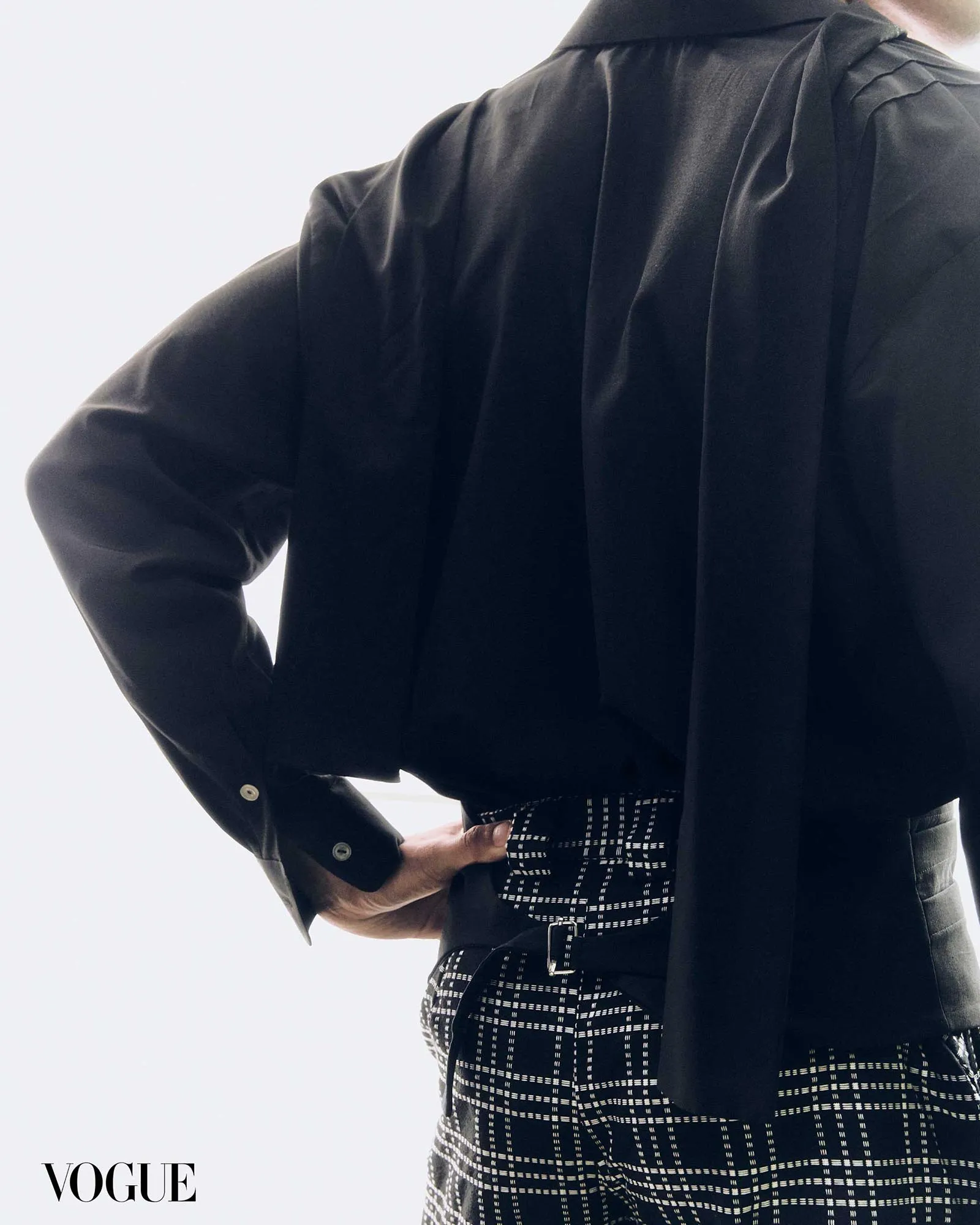 Close-up of a person wearing a black blouse over a black-and-white checkered garment, with a Vogue logo in the bottom-left corner.