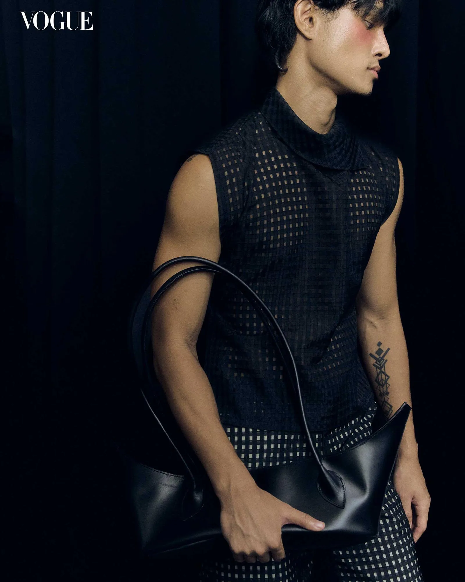 Profile of a fashion model in a black, grid-pattern sleeveless top, holding a black bag; visible geometric tattoo on forearm, Vogue logo in corner.