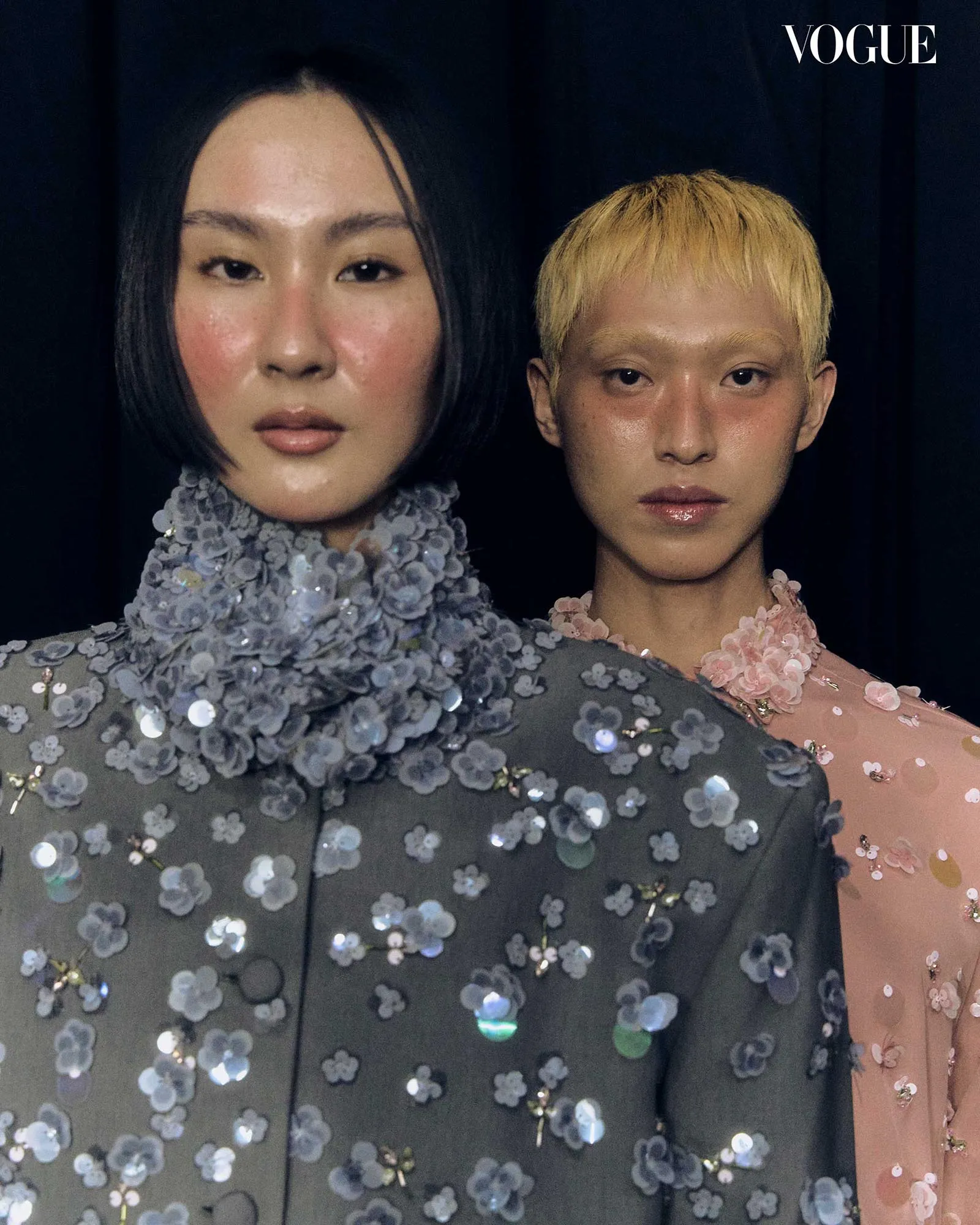 Two models pose closely in sequined gowns against a dark background, Vogue logo in the top-right corner.