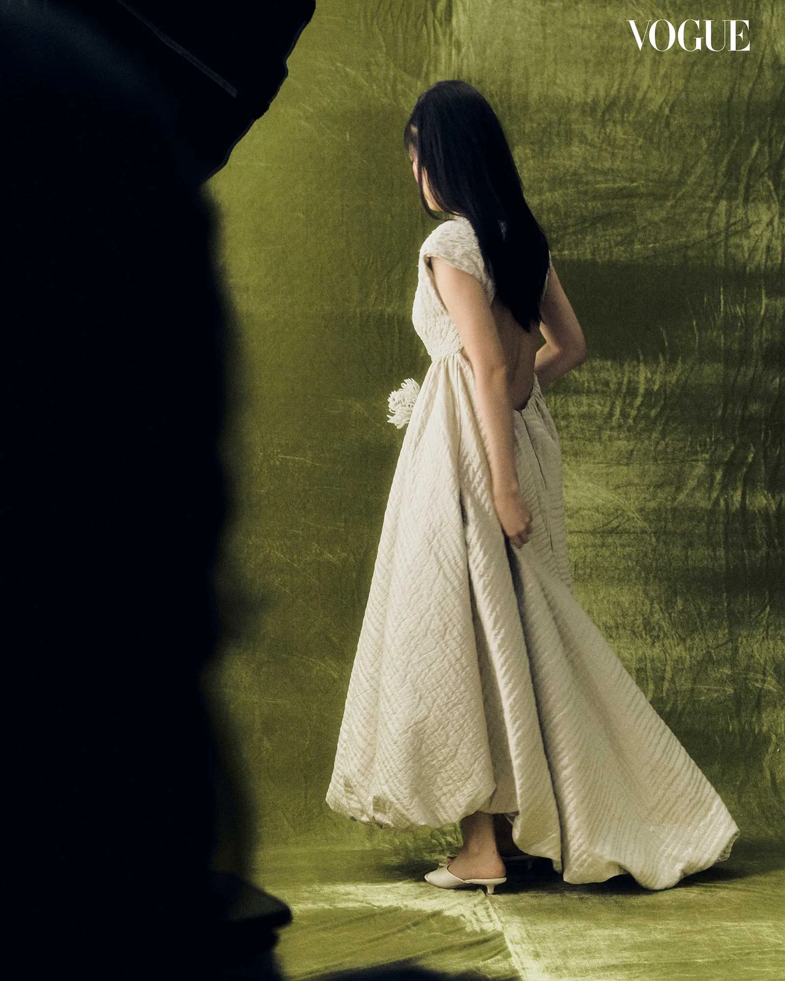 Side view of a woman in a cream textured dress walking on a gold studio backdrop, holding a small white bouquet; Vogue logo in the corner.