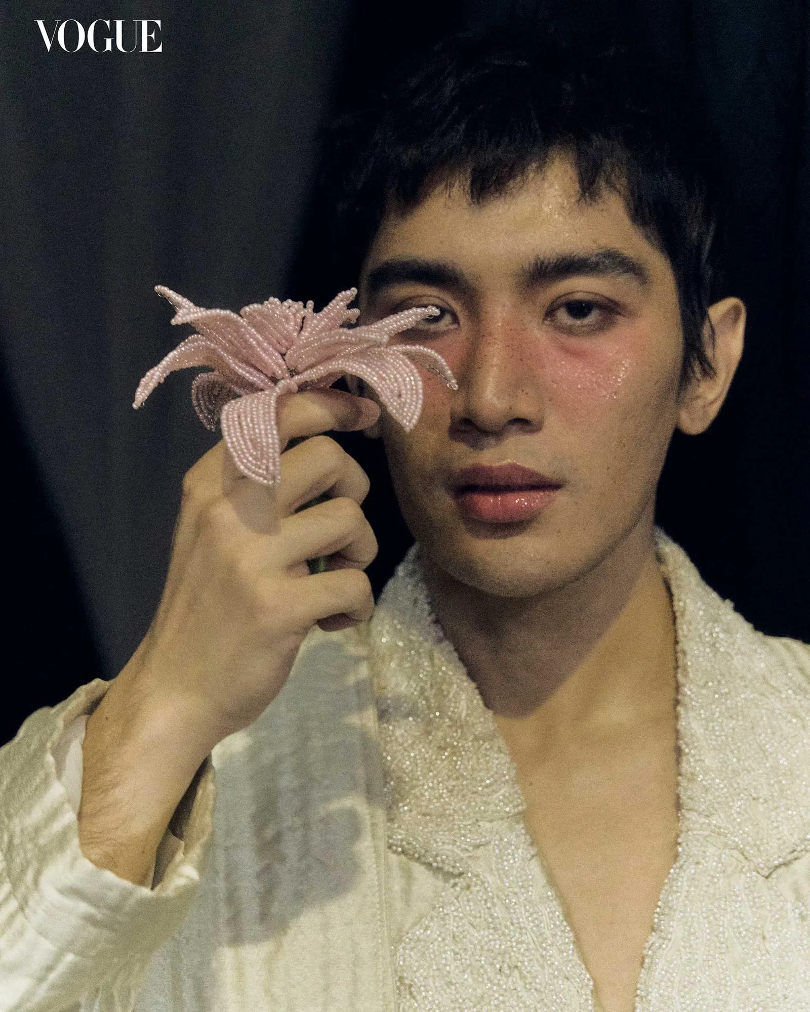 Close-up portrait of a person with short dark hair wearing a shimmering white outfit, holding a pink beaded starfish near the eye; Vogue logo is in the top-left corner.
