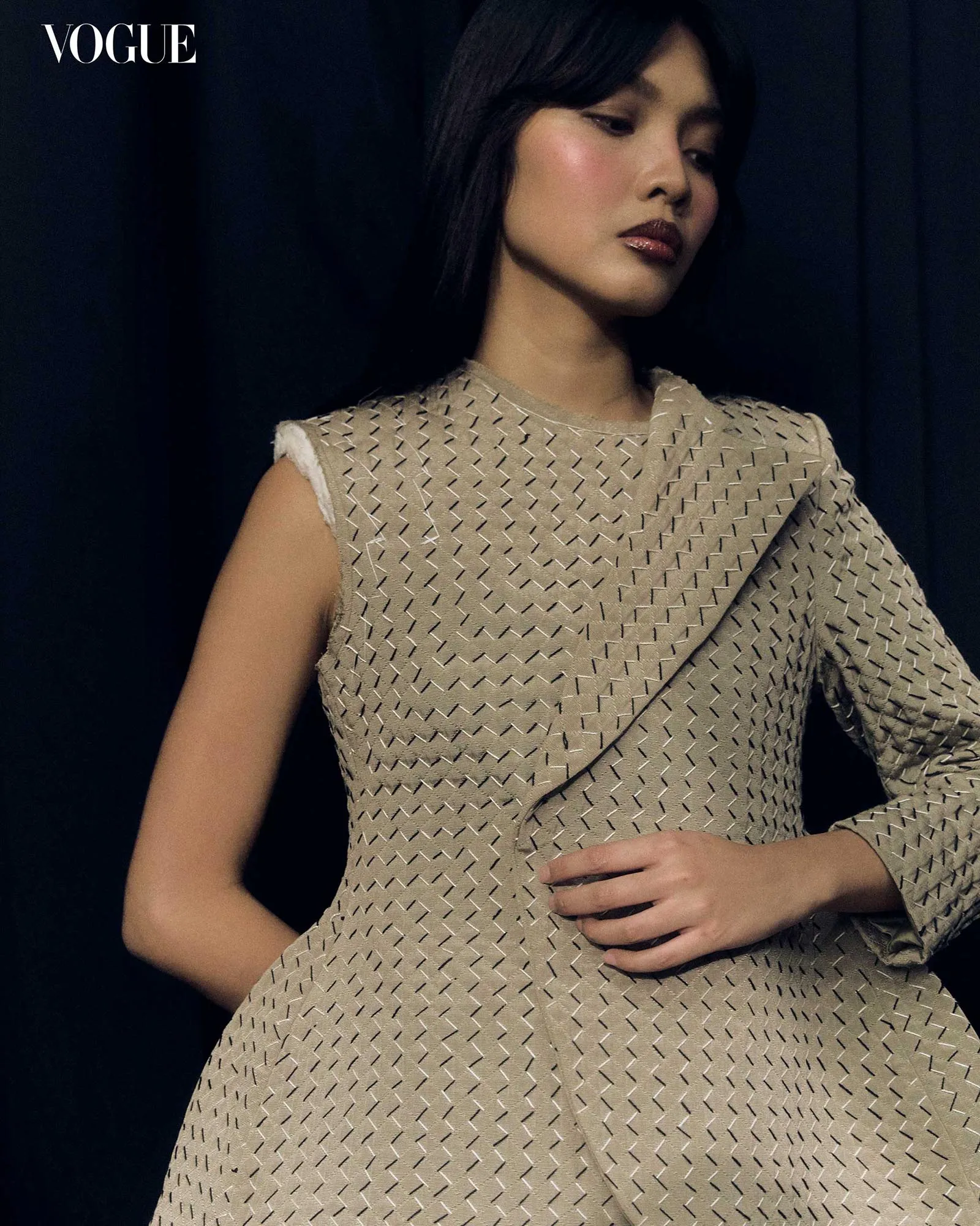 Model in a beige textured dress poses against a dark backdrop with a Vogue logo in the corner.