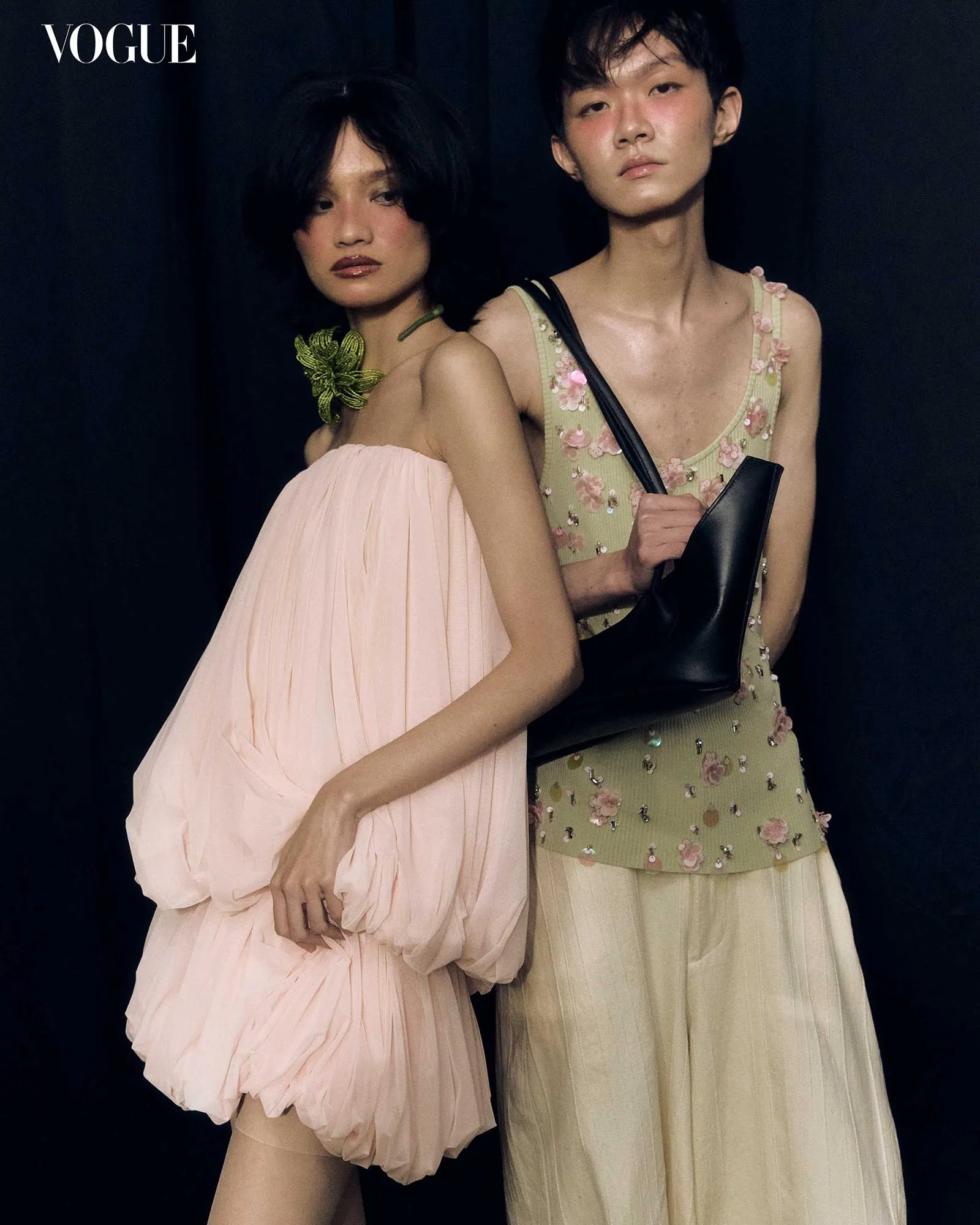 Two fashion models pose side by side against a dark backdrop; left in a pale pink off-shoulder dress with voluminous sleeves and a green floral necklace, right in a pale green floral dress holding a black handbag strap.