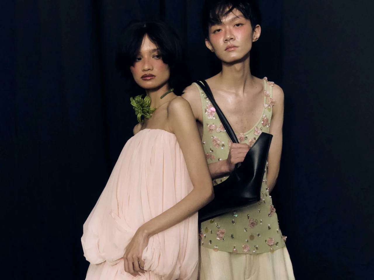 Group of five models posing backstage in fashion outfits, with three women in embellished pink and beige dresses and two men in light-toned attire against a dark backdrop.