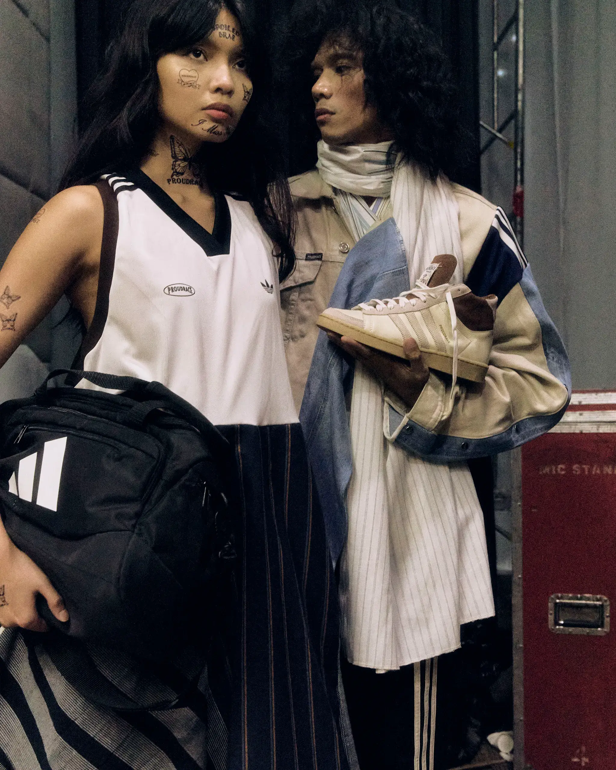 Two tattooed individuals posing closely; one holds a beige sneaker in front of a dark backdrop, wearing striped clothing and a bag in view.