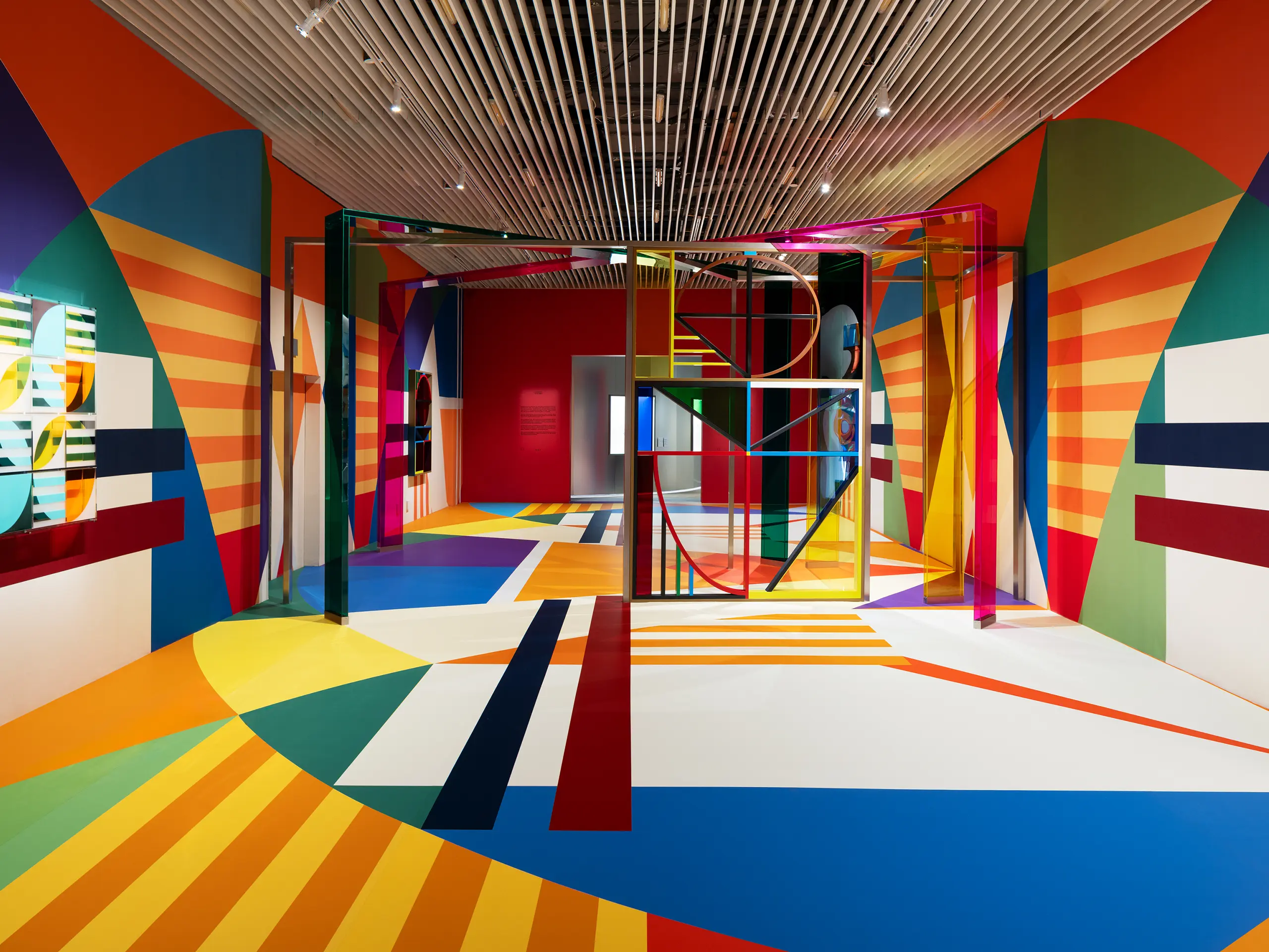 Vibrant abstract gallery interior with rainbow-striped walls and floor, and a glass-framed sculpture in the center.