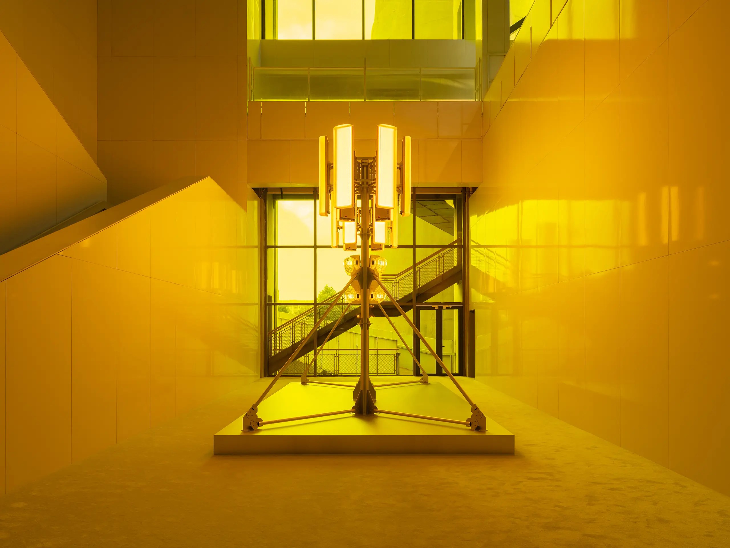 Centerpiece lighting sculpture on a pedestal in a yellow-lit modern atrium with glass walls behind it.