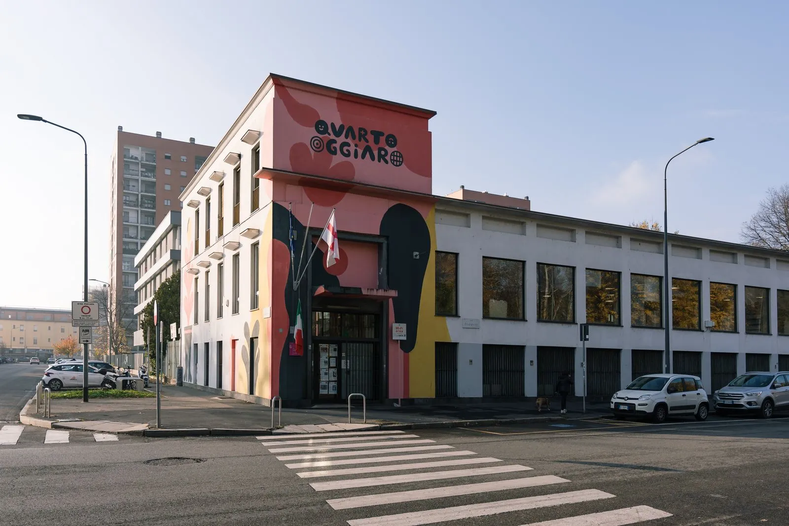 Colorful mural-covered building on a street corner with a crosswalk in the foreground and parked cars along the curb.