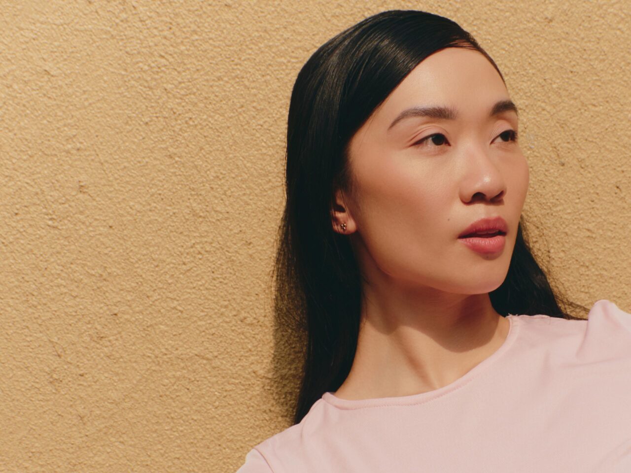 Close-up portrait of a woman with dark hair looking to the right, against a beige textured wall.