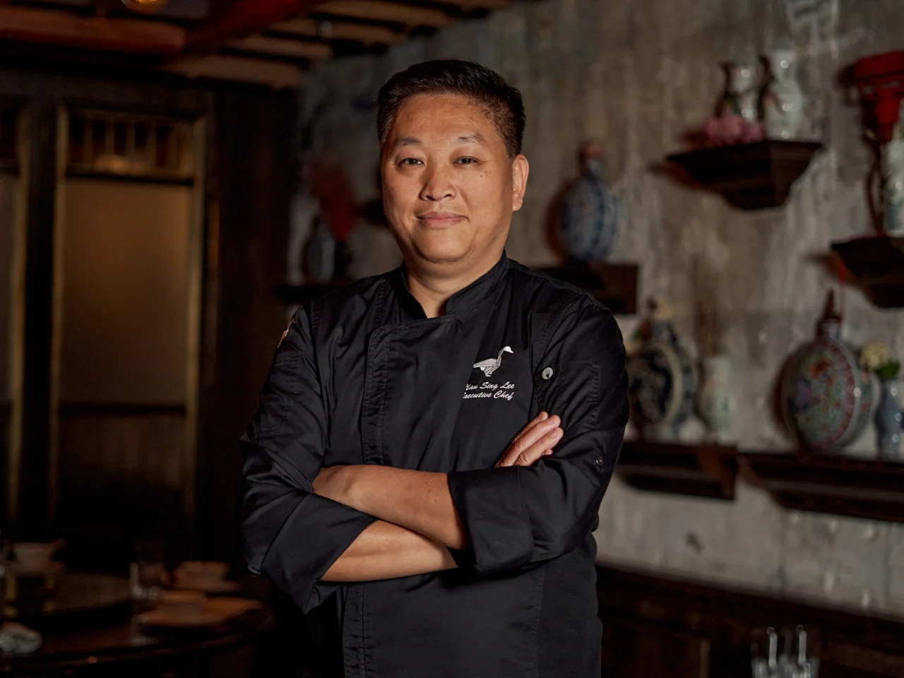 Professional chef in a black uniform stands with arms crossed in a dimly lit kitchen, decorative ceramics on shelves in the background.