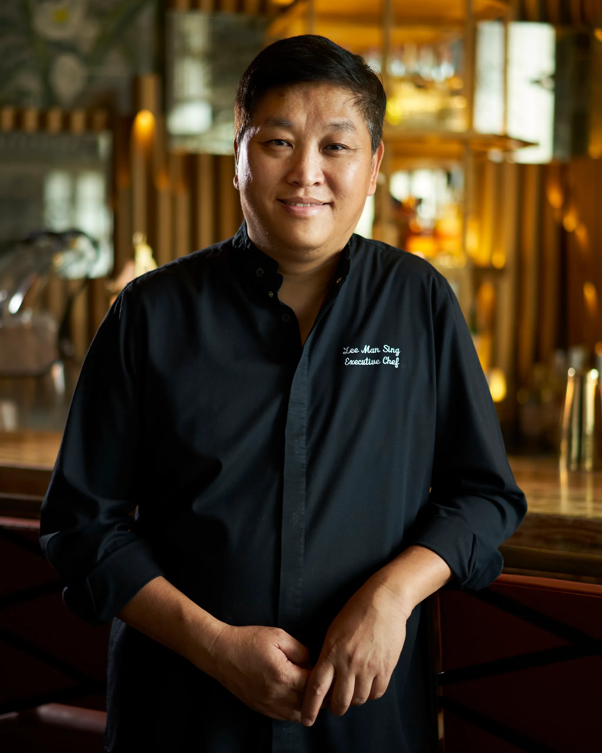 Portrait of Chef Lee Man Sing in a black chef's coat, smiling, in a warmly lit restaurant backdrop and wooden decor.