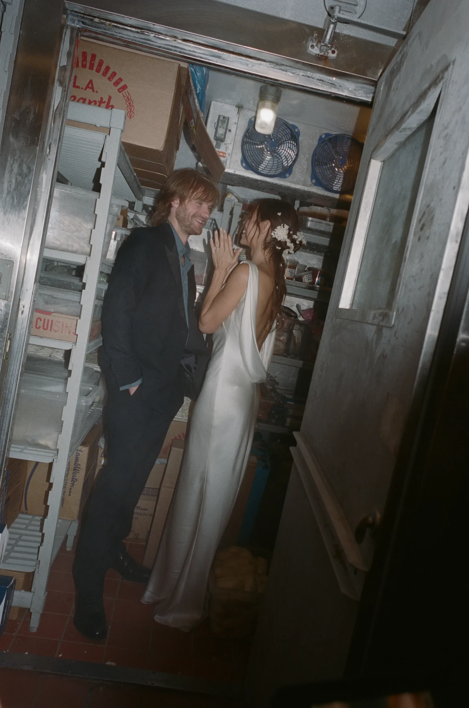 Bride in a backless white dress with a floral hairpiece and a groom in a dark suit share a kiss in a cluttered industrial storage room with metal shelves and boxes nearby.