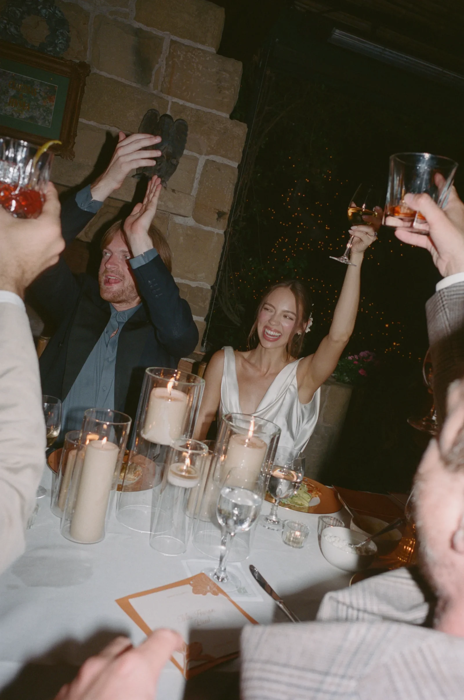 People at a dinner party raise glasses in a toast around a candlelit table with a stone wall backdrop.