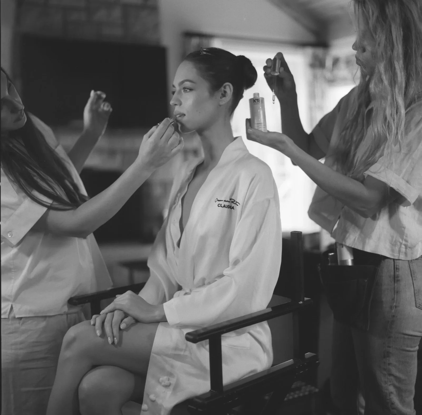 Makeup artist applies lipstick to a seated woman while another stylist holds a spray can nearby in a backstage studio scene, seating her for touch-ups.