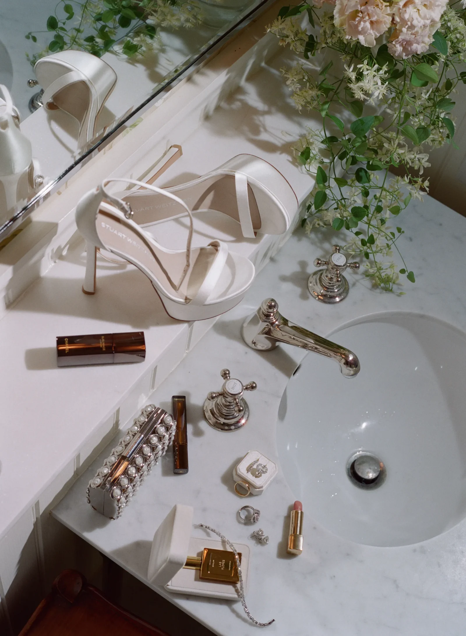 White strappy high-heeled sandals on a marble bathroom counter beside jewelry and cosmetics, with a mirror and greenery in the background.