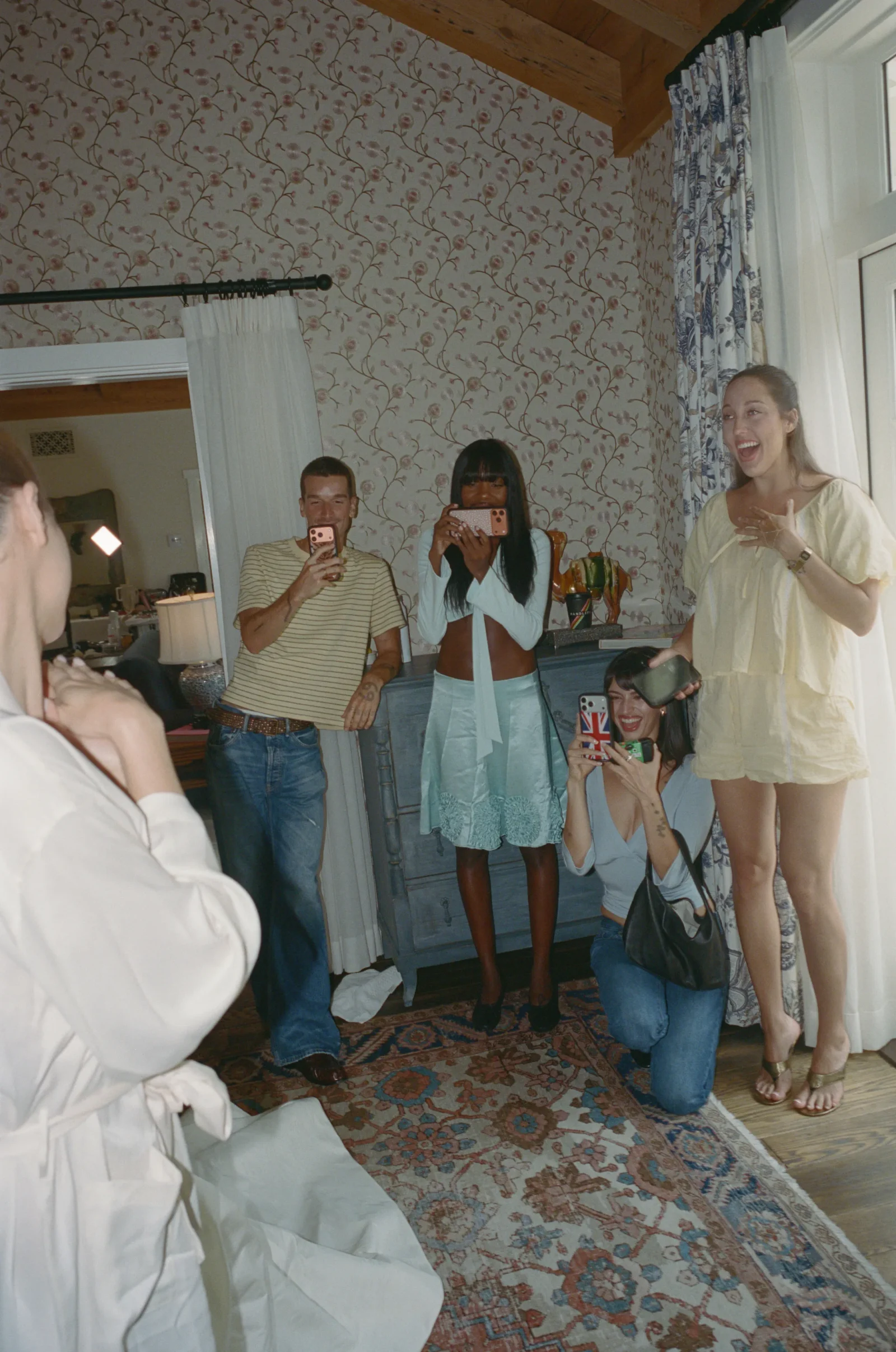 Group of friends posing for a mirror selfie at a cozy home party, phones raised and smiling.