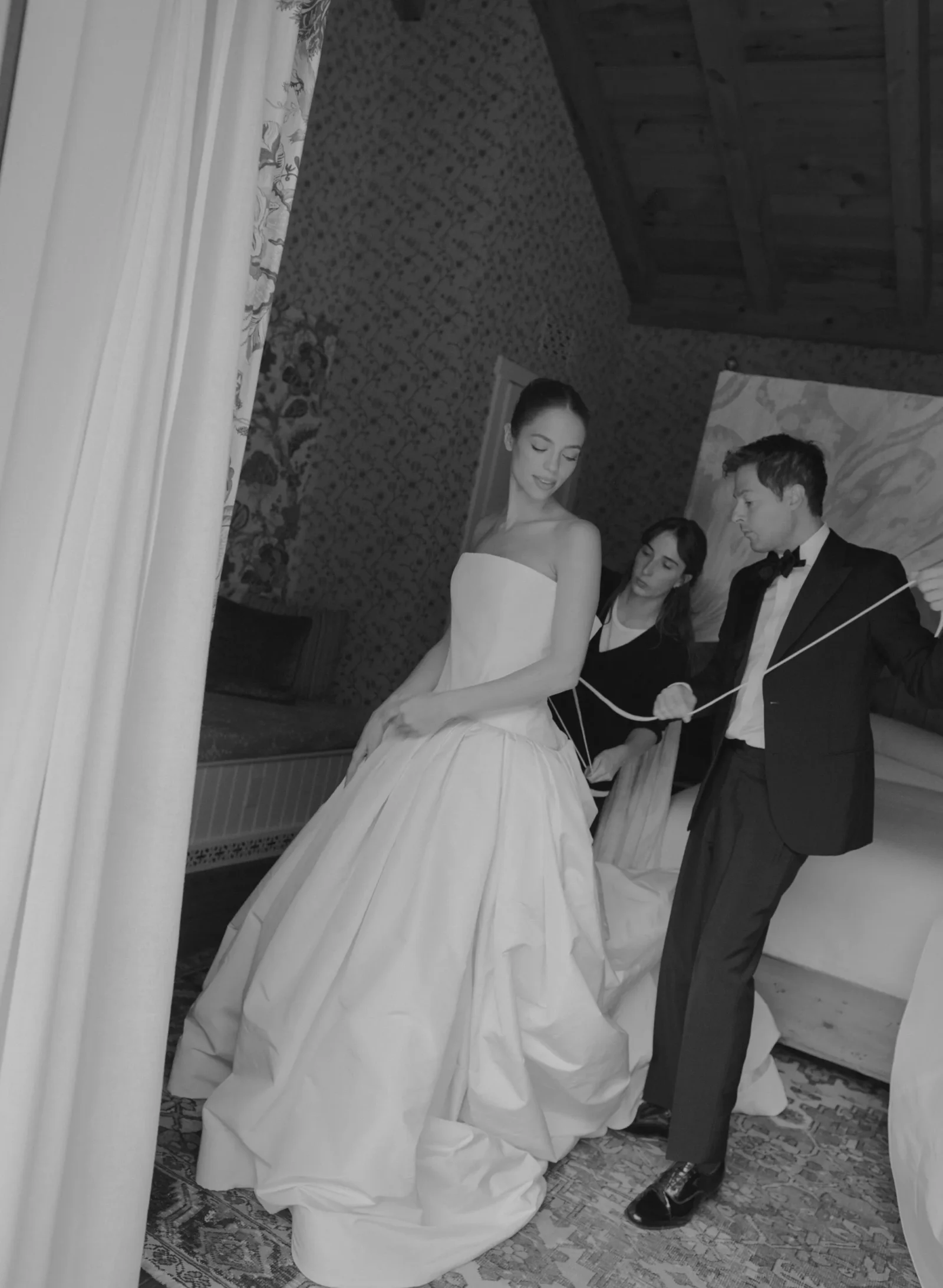Bride in a strapless white wedding gown stands as two assistants and a man in a tuxedo help adjust her dress in a room with patterned wallpaper and curtains.