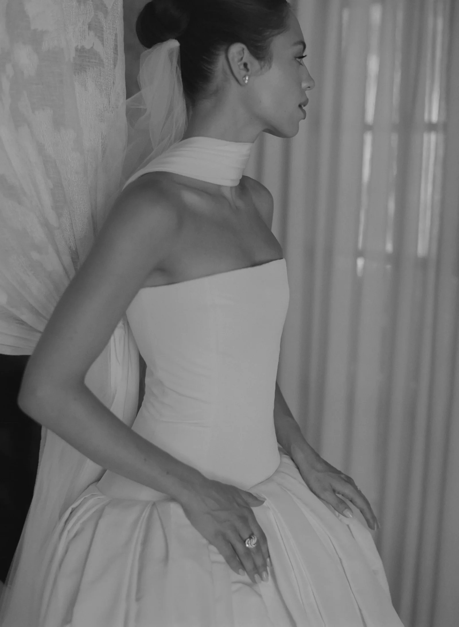 Bride seated in profile wearing a white strapless gown and veil, with an assistant adjusting the dress behind her.