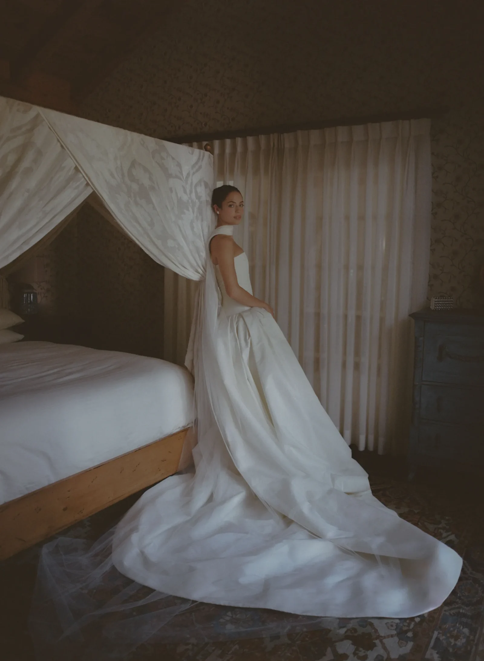 Woman in a white wedding gown sits on a bed under a lace canopy in a softly lit bedroom.