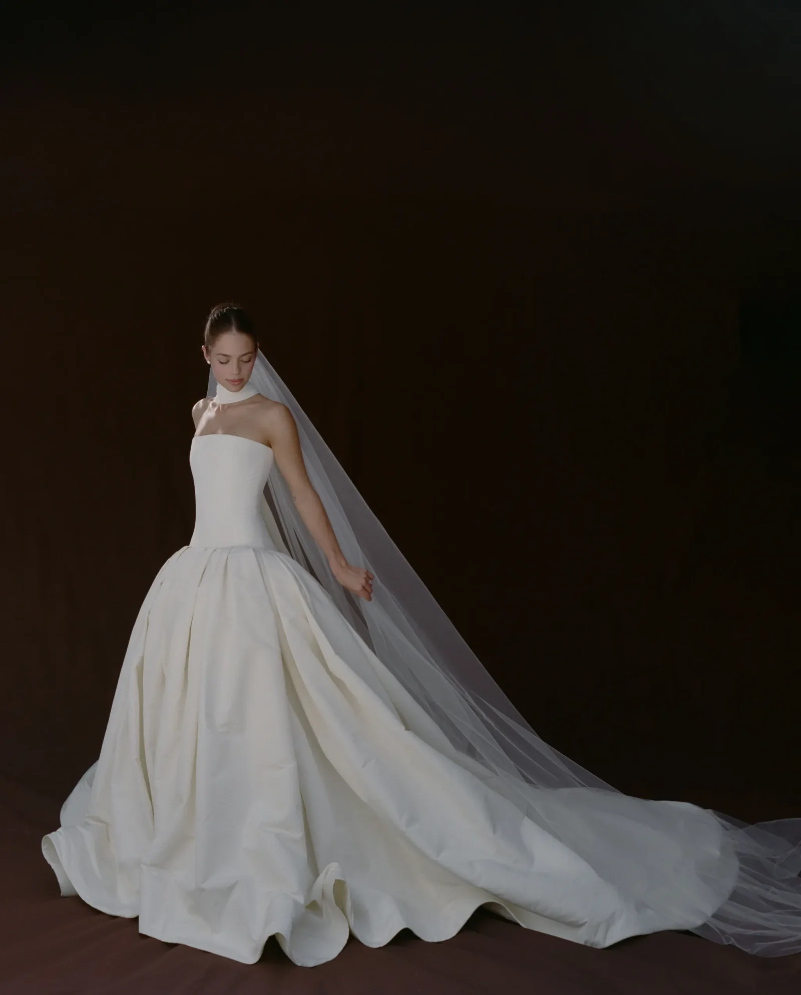 Bride in a strapless white wedding gown with a long veil flowing behind her against a dark backdrop, lower left corner of the frame.