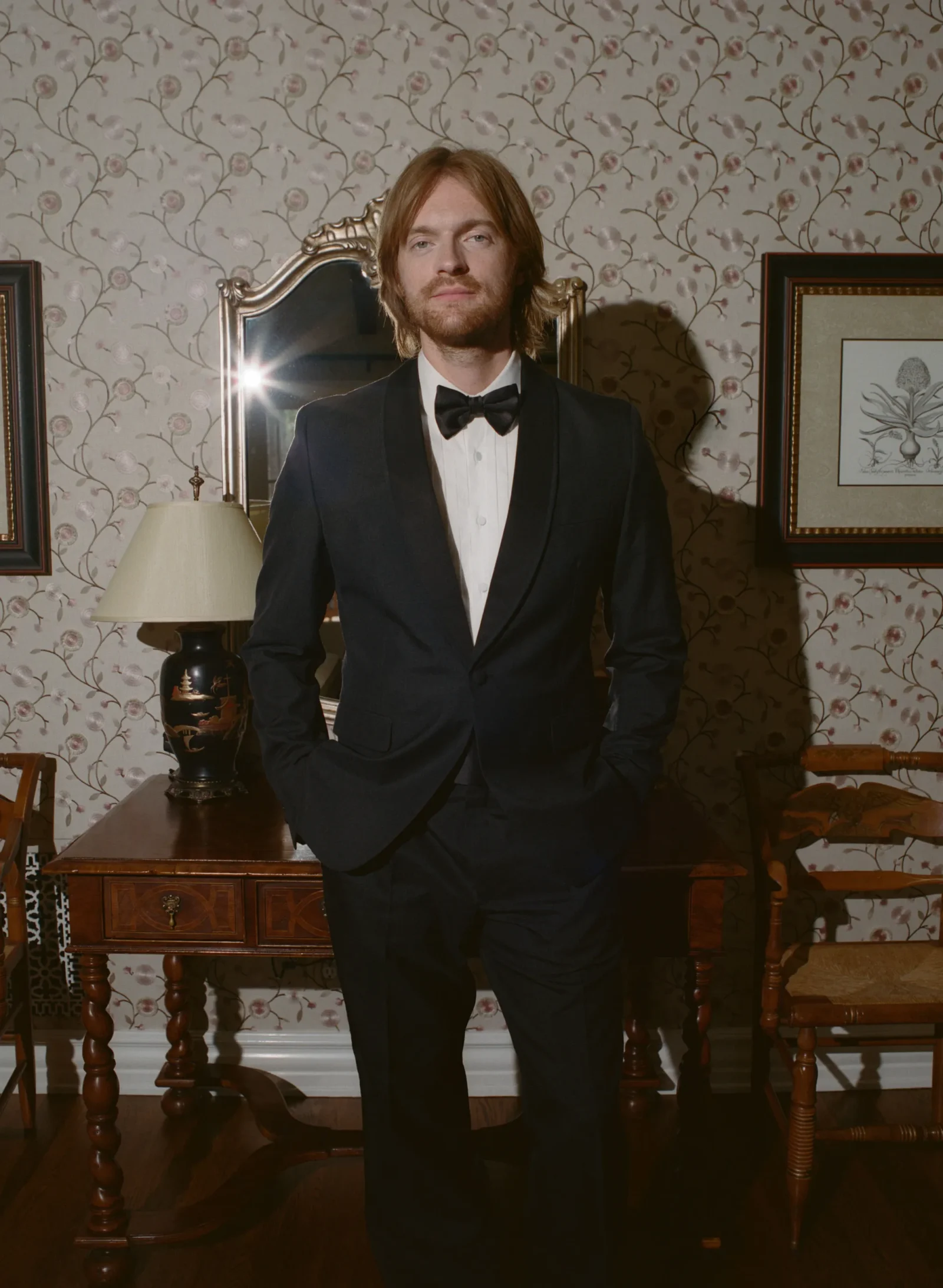 Young man in a black tuxedo and bow tie stands indoors in a vintage room with floral wallpaper.