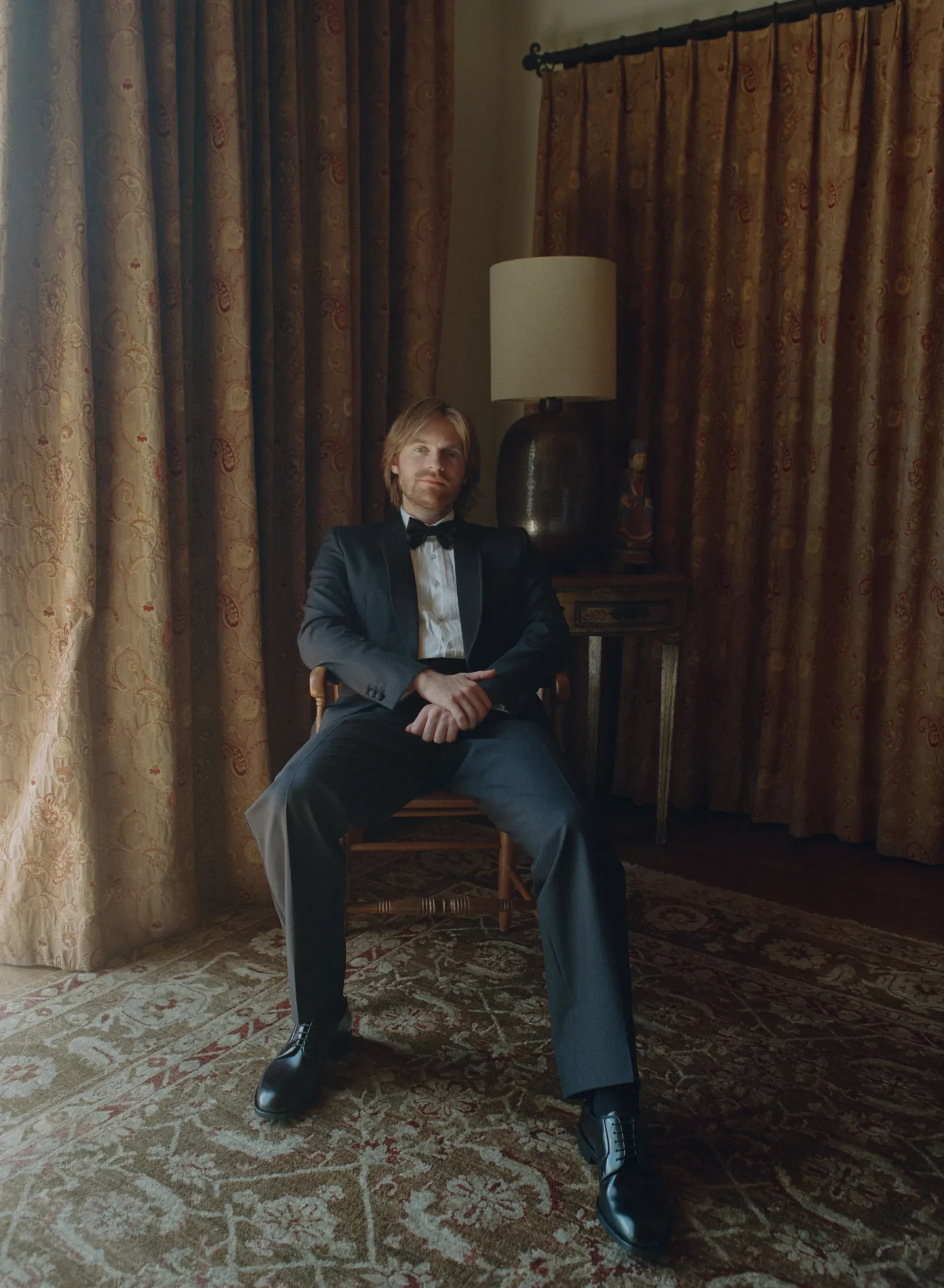 Man in a dark suit and bow tie sits in a wooden chair in a vintage living room with heavy patterned curtains and a lamp behind him.