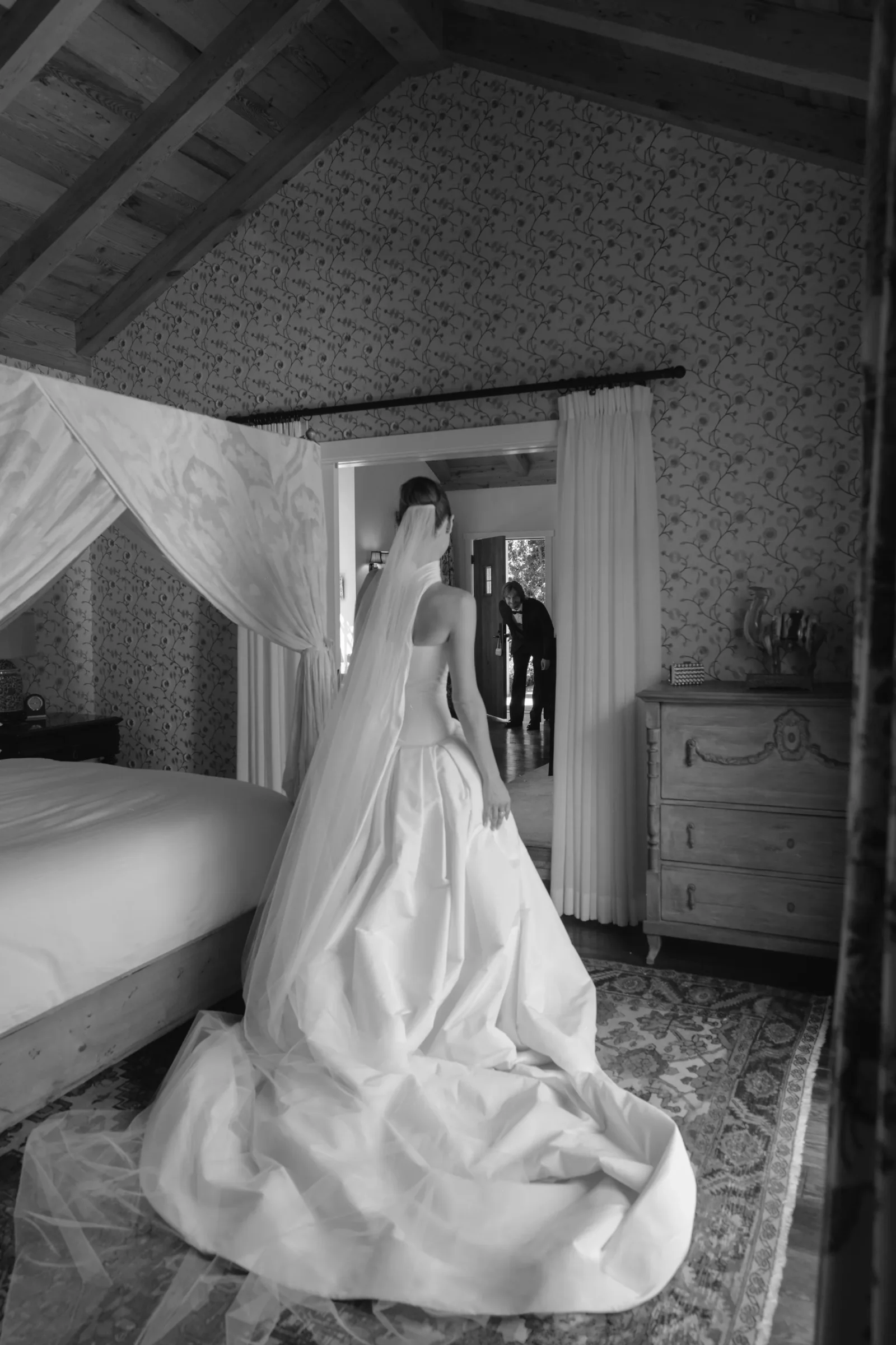 Bride in a strapless wedding gown walks through a rustic bedroom with exposed wooden beams and a canopy bed, patterned wallpaper in the background.