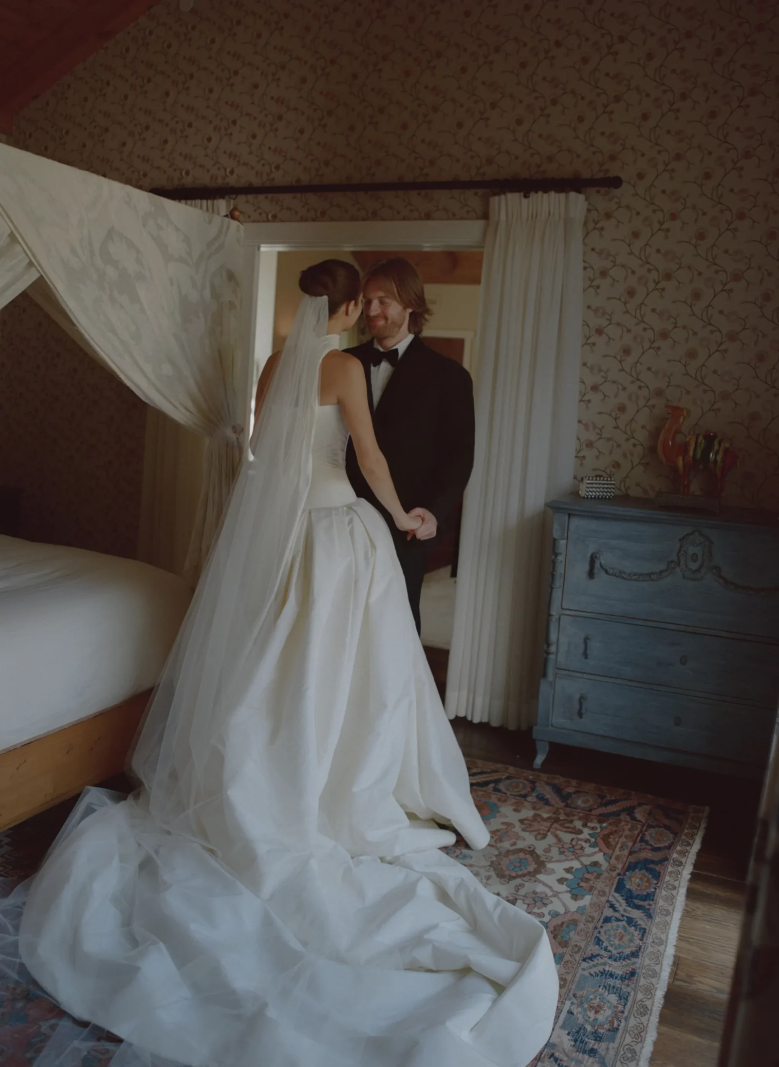 Bride in a white wedding gown and veil stands with her groom in a black tuxedo inside a vintage bedroom with floral wallpaper and draped curtains.