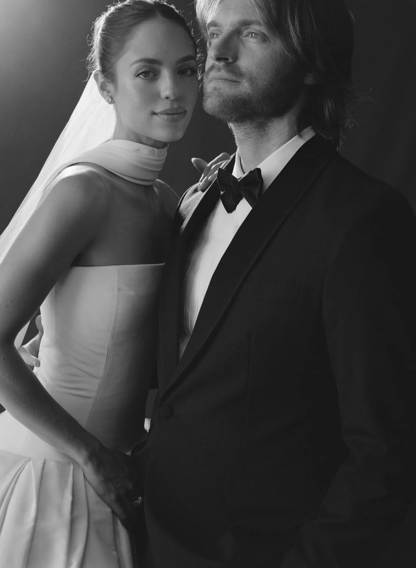 Black-and-white photo of a bride and groom posing together; she wears a strapless dress with a veil, he in a tuxedo with a bow tie, both looking at the camera with calm expressions.