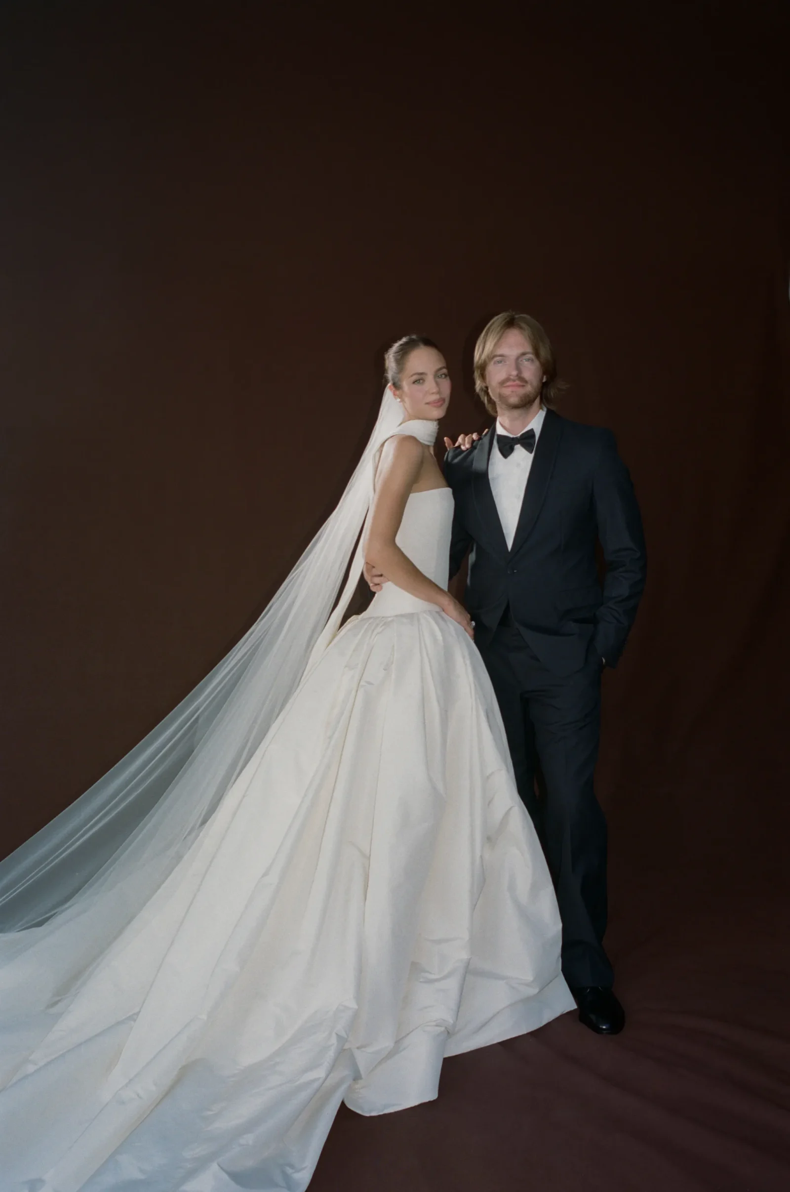 Bride in a white wedding gown and long veil posing with a groom in a black tuxedo against a dark brown backdrop, both looking at the camera.