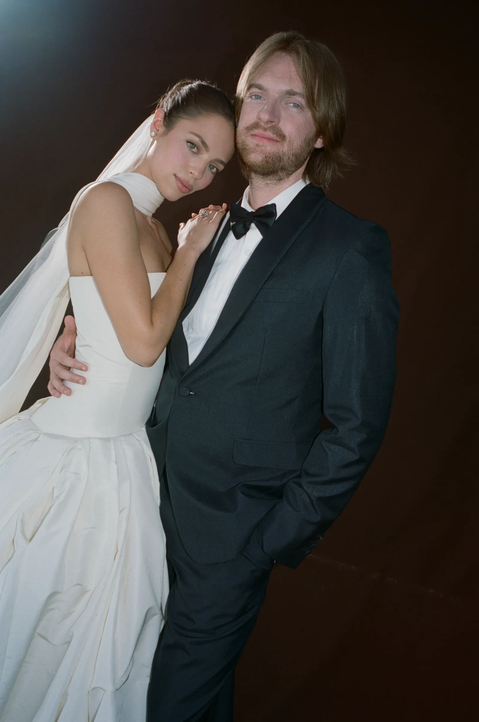 Bride in a white gown and veil rests her head on a groom's shoulder as they pose closely in a dark studio background, wedding-style portrait.