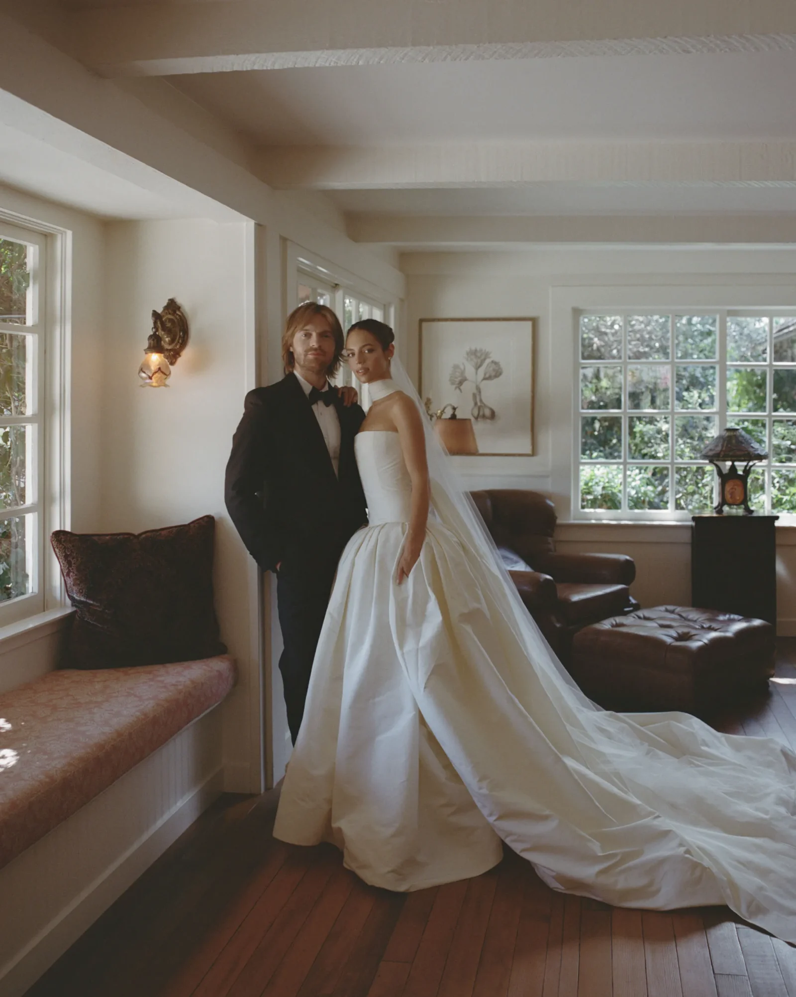 Bride in a white gown and groom in a black tuxedo pose together in a bright living room interior, wedding photo.**
