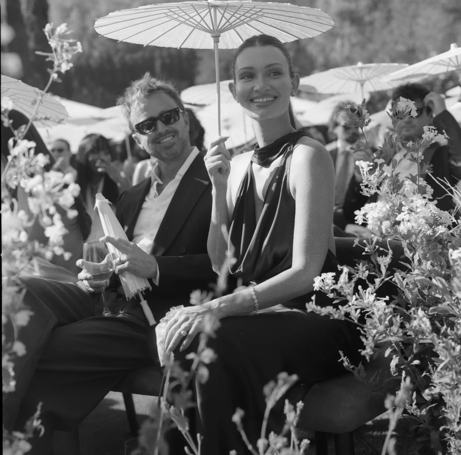 Woman smiling under a paper parasol at an outdoor garden party, with guests and flowers in the background.