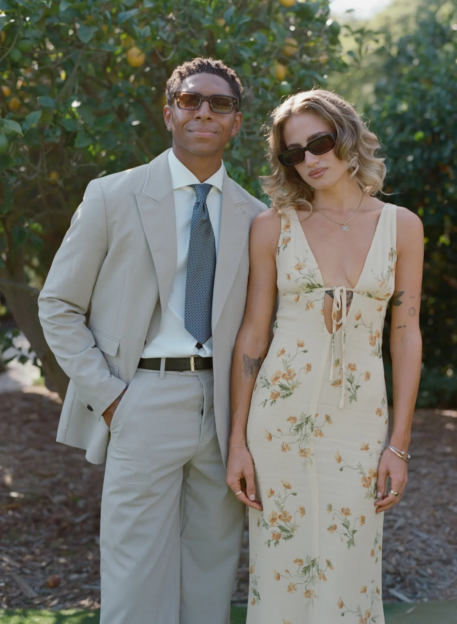 Two people posing together outdoors in a sunny garden; a man in a light gray suit and tie beside a woman in a cream floral dress, both wearing sunglasses.