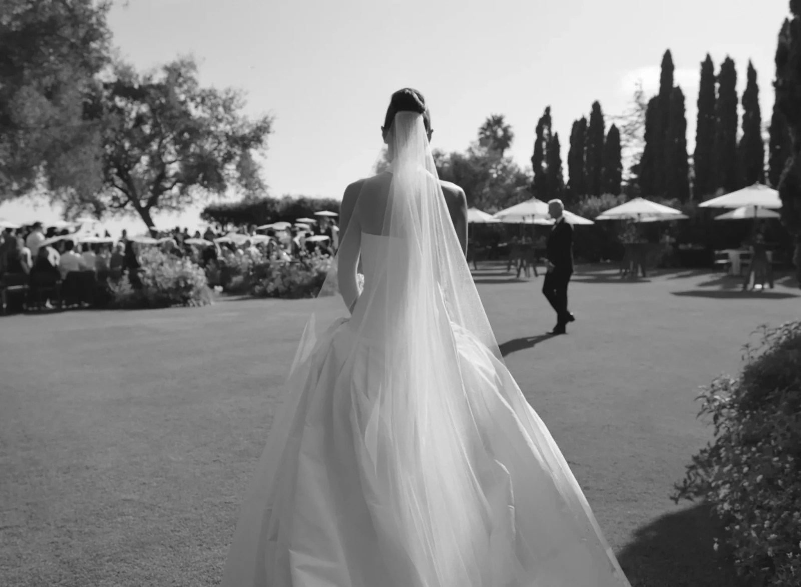 Bride in a wedding dress with veil walking away outdoors at a reception, guests seated under umbrellas in a garden setting
