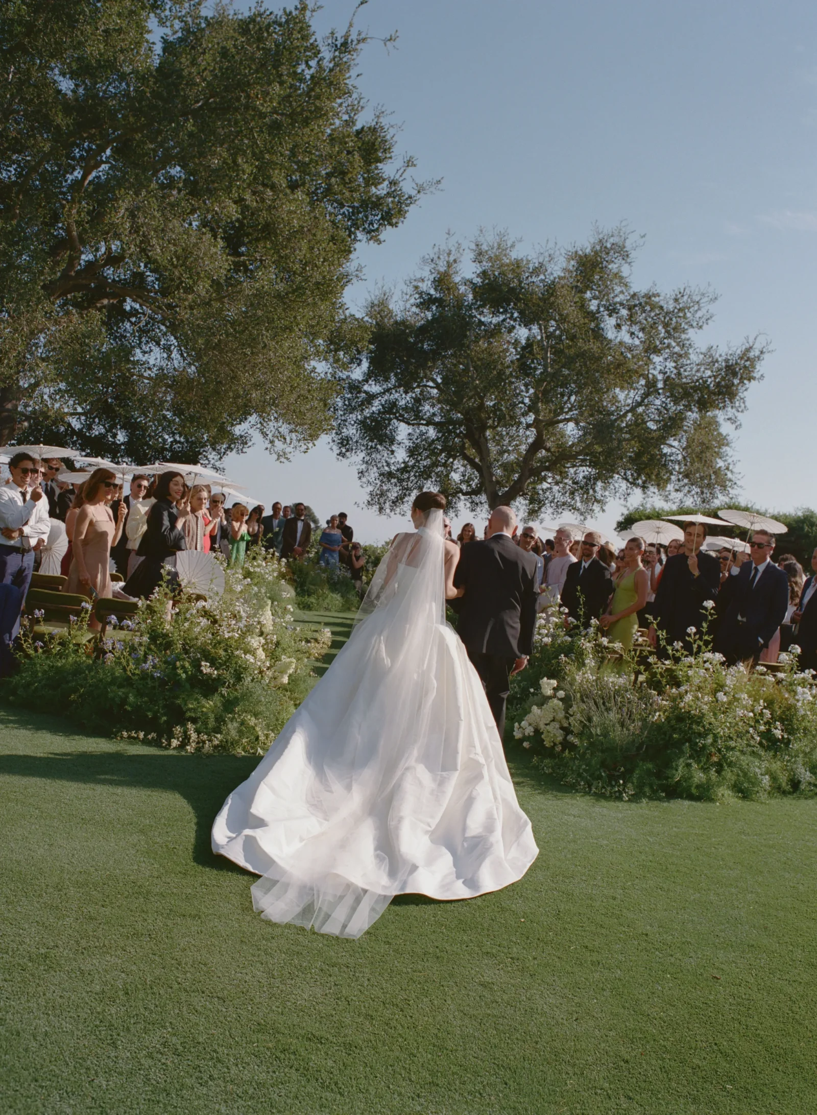 Bride and groom walk down a garden aisle as guests stand with white parasols under trees on a sunny outdoor wedding. The bride wears a long white gown with a veil.