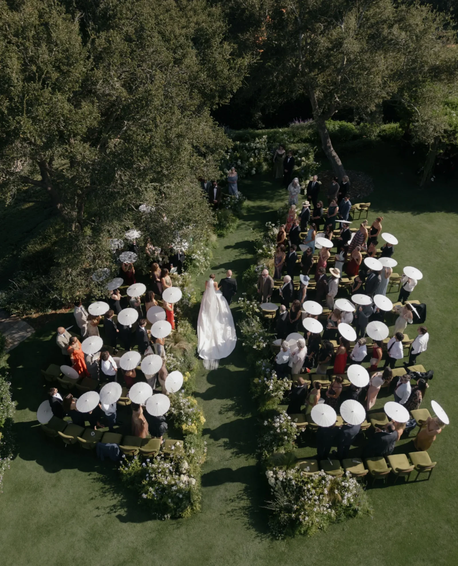 Aerial view of an outdoor wedding ceremony with guests seated in rows under white parasols on a green lawn amid trees and flowers; the bride in a white gown stands near the center.