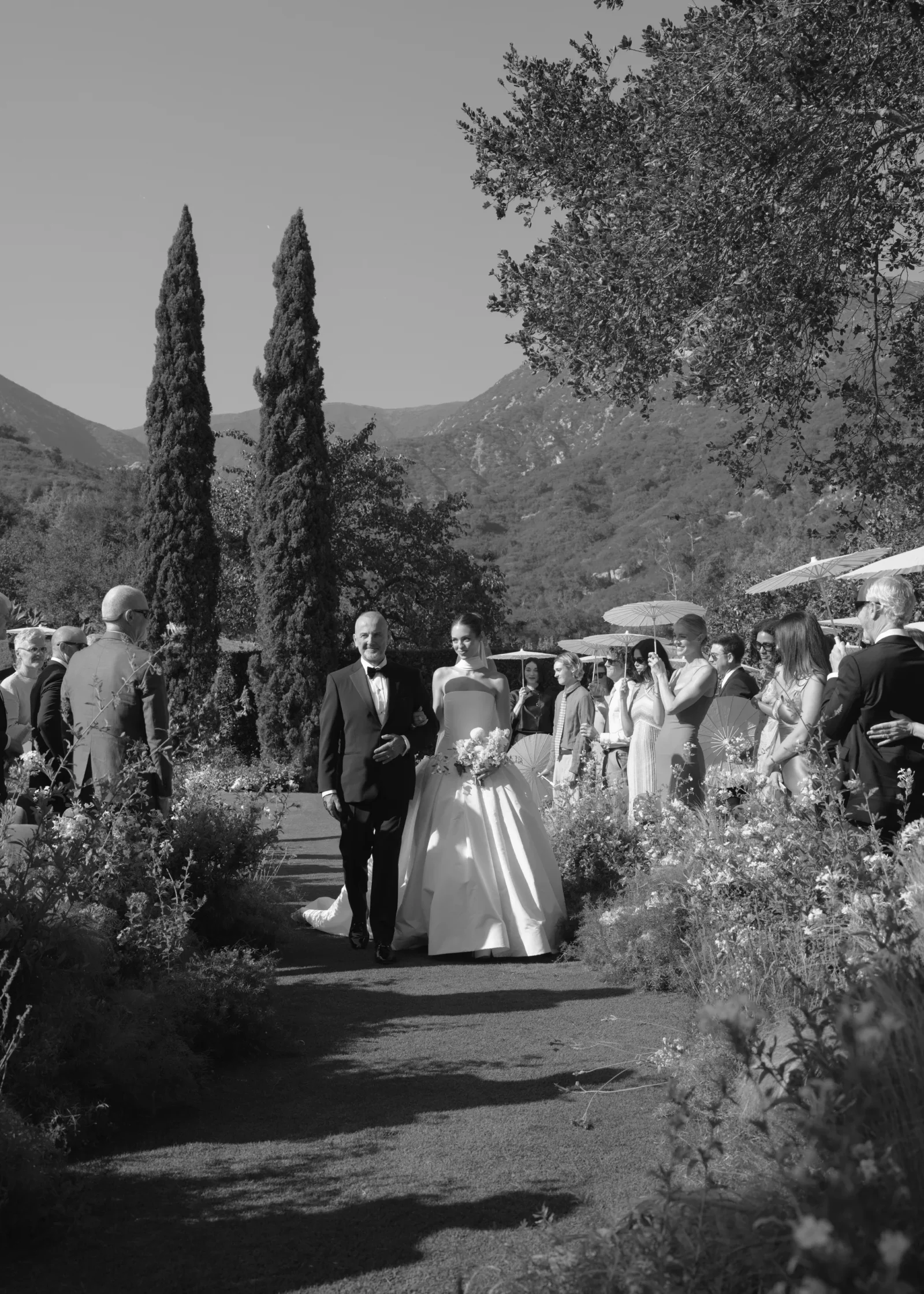 Bride in a ball gown and groom in a tuxedo walk down a garden aisle as guests stand along the path in an outdoor wedding setting, mountains in the background (black and white).