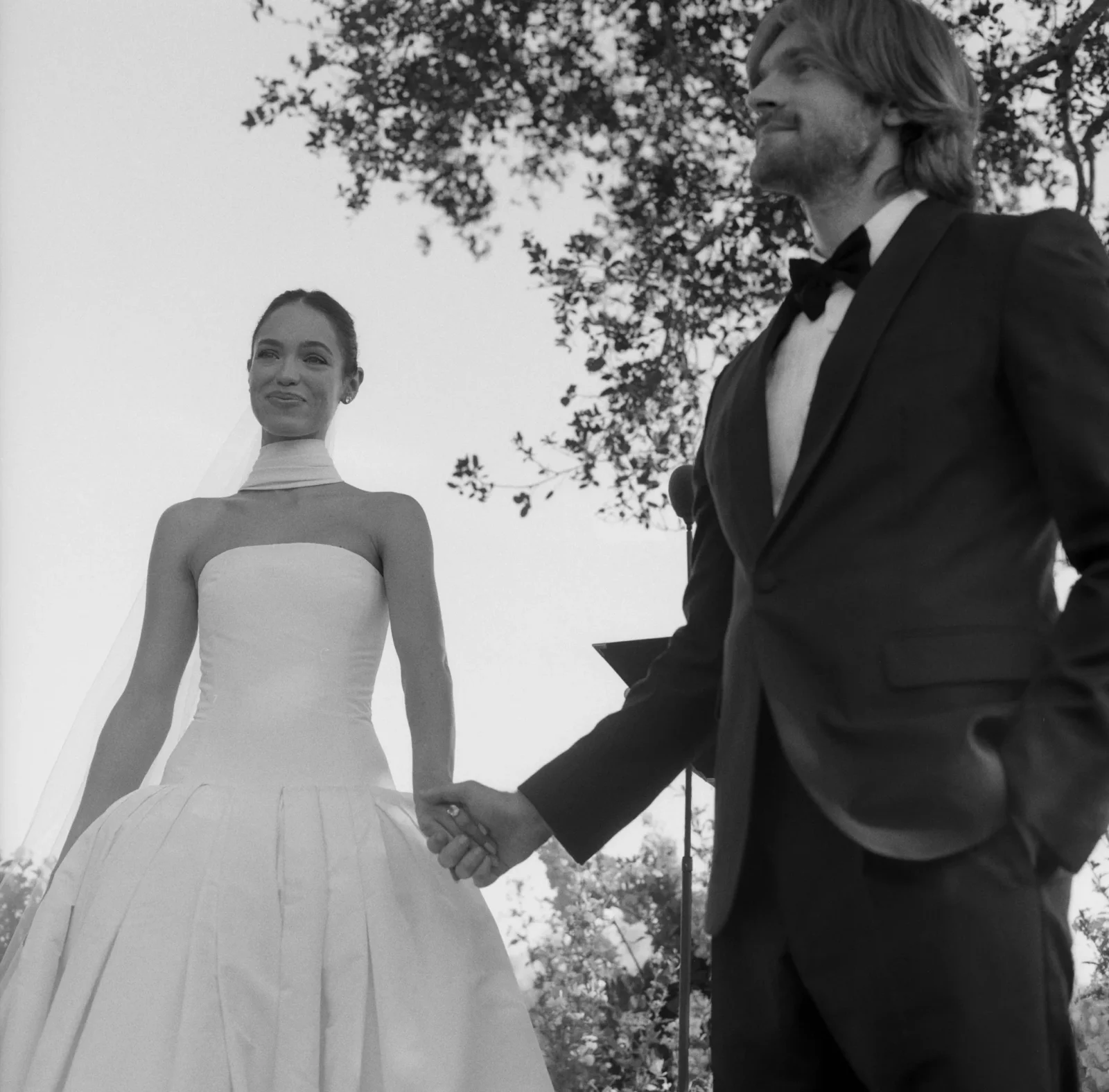 Bride in a strapless wedding dress and veil holds hands with a man in a dark suit outdoors, with trees in the background.