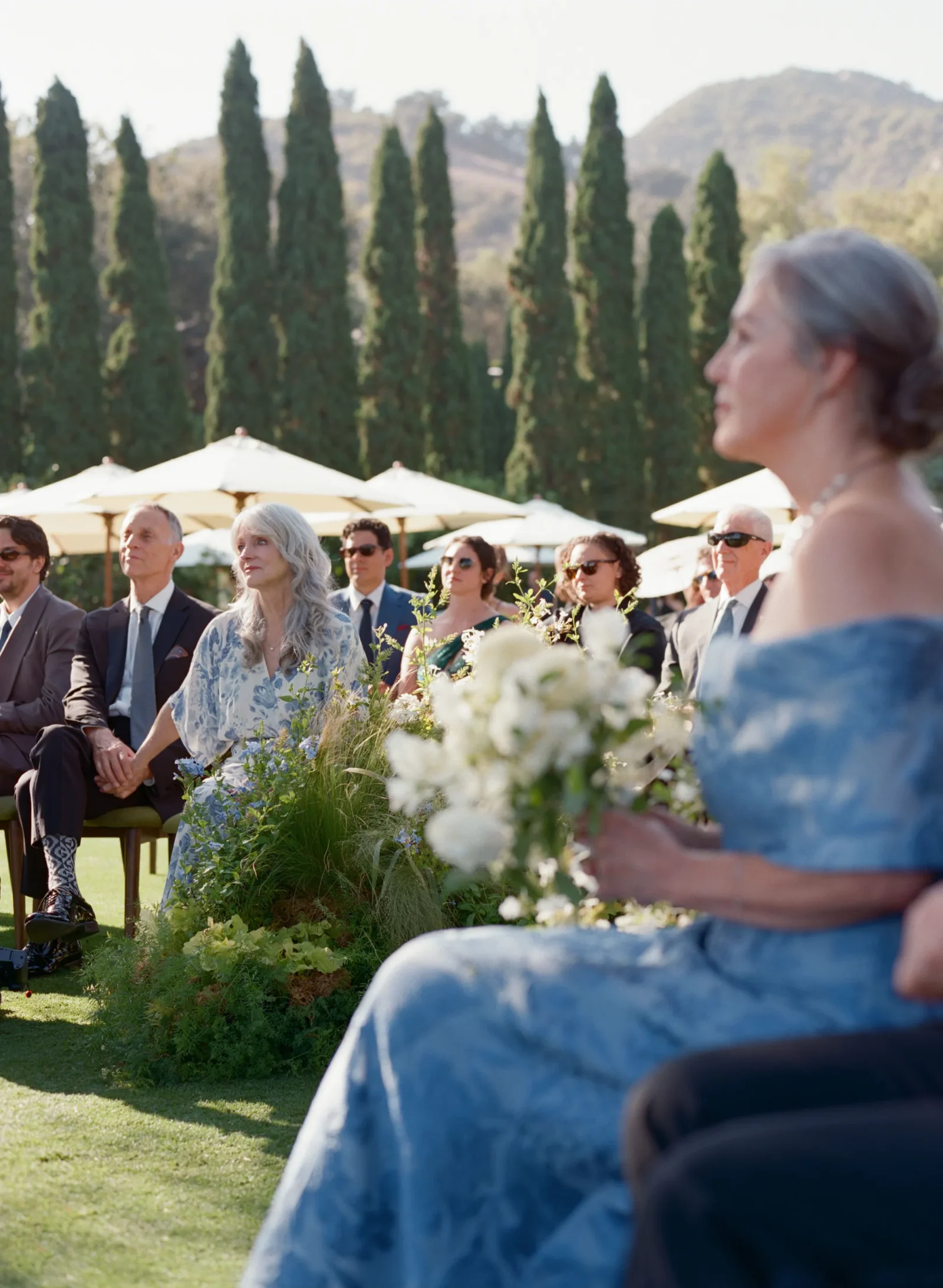Outdoor wedding ceremony with guests seated in formal attire under white umbrellas and lush greenery, hills in the background.