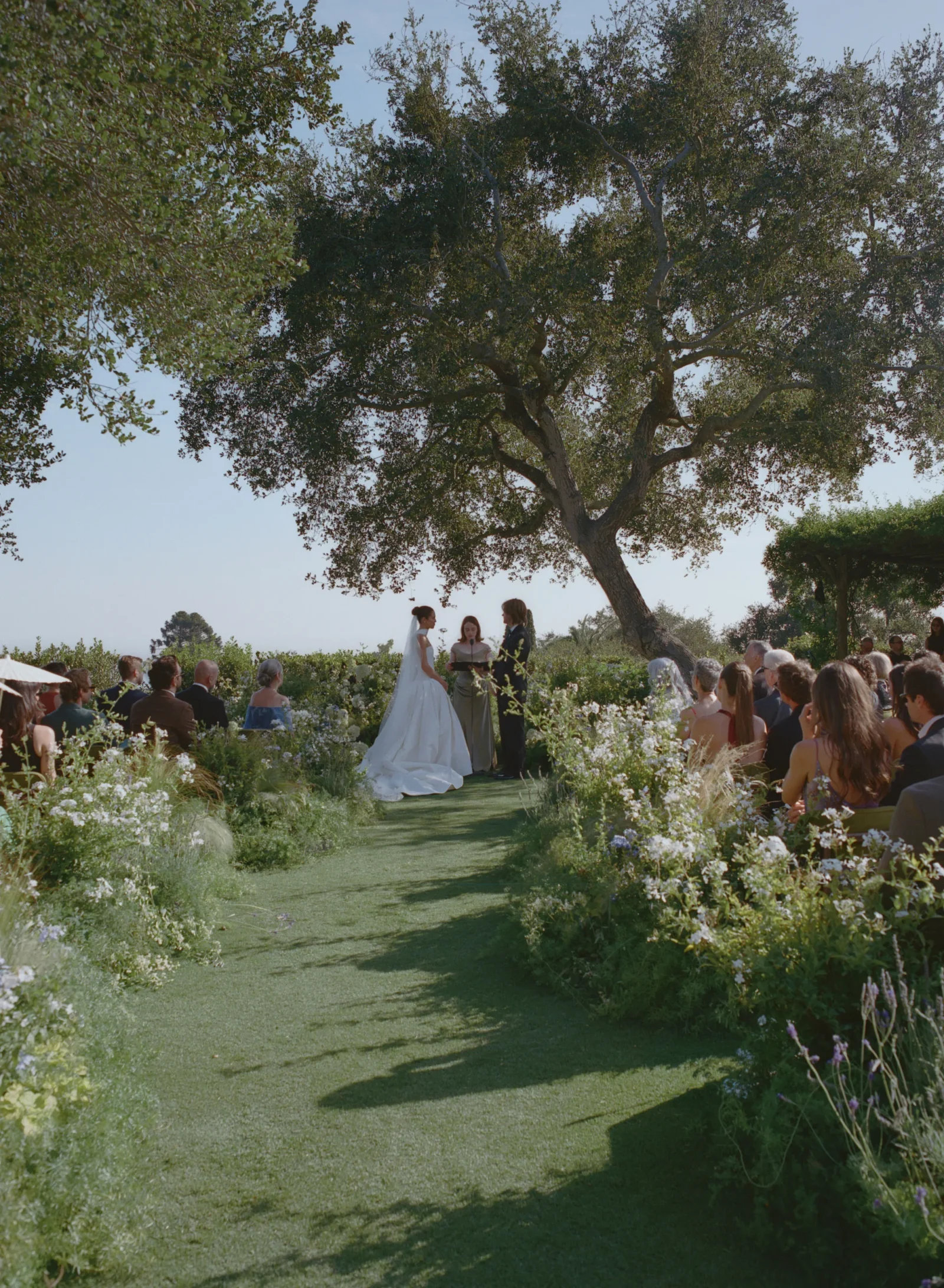 Bride and groom exchange vows beneath a large tree at an outdoor wedding, officiant reading as guests sit along a flower-lined aisle.
