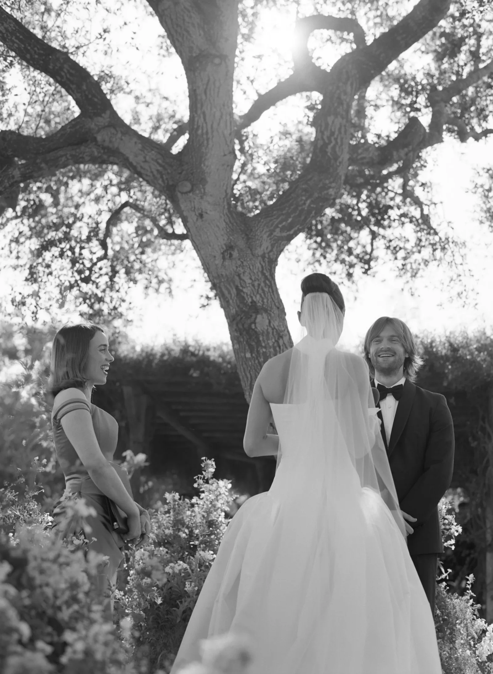 Bride in a white wedding gown with veil standing next to a smiling groom in a suit, outdoors beneath a large sunlit tree.