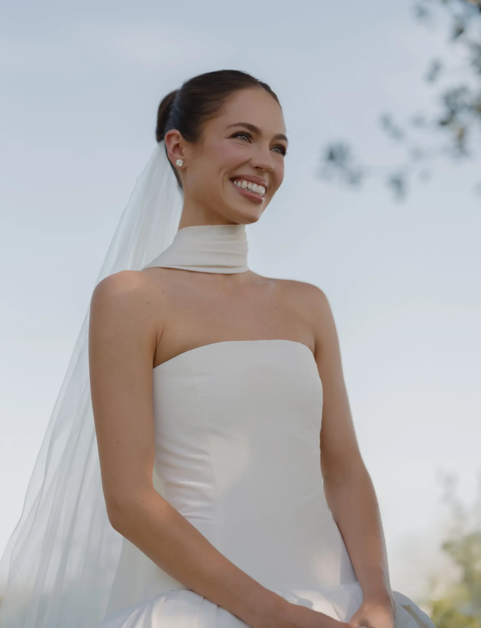 Bride in a strapless white wedding gown with a veil, smiling outdoors against a clear blue sky.