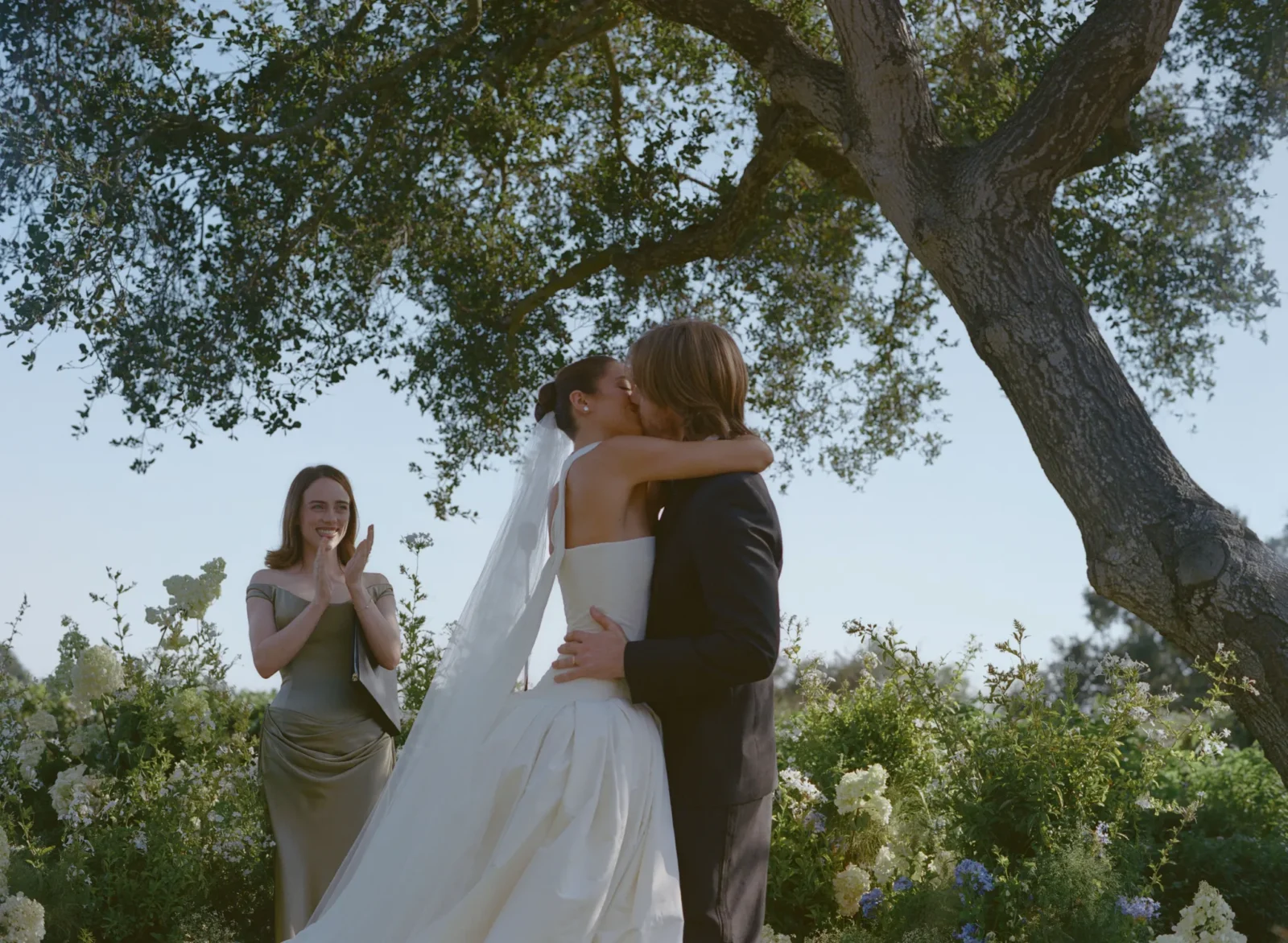 Bride and groom kiss in an outdoor garden setting, a bridesmaid claps nearby among white flowers and greenery.